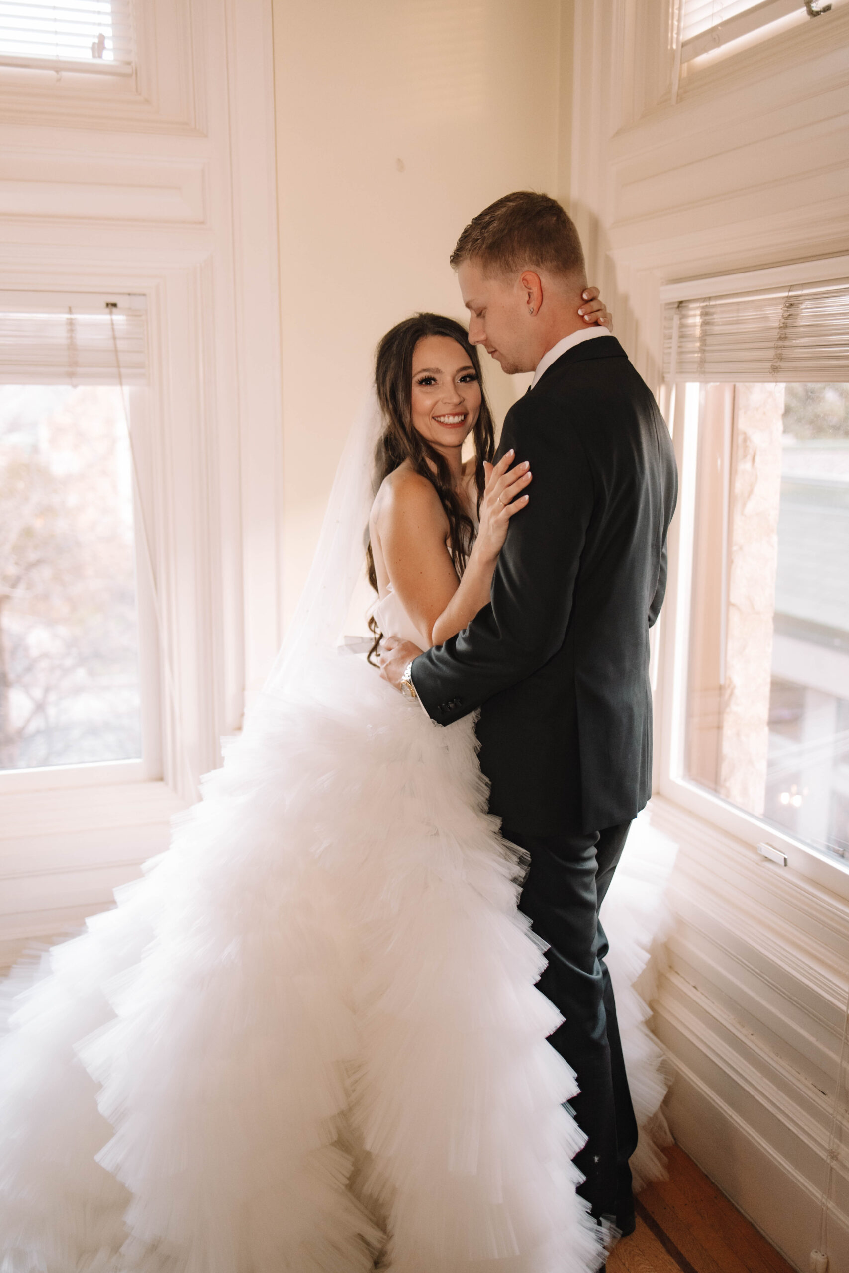 A bride in a white gown and a groom in a black suit share an intimate moment by a window in a softly lit room.