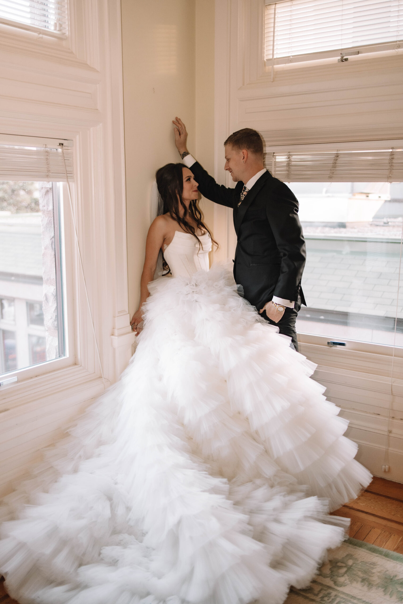 A bride in a voluminous white gown leans against the wall as her husband leans in for a kiss. Her dress from a wedding dress shop in Minneapolis 