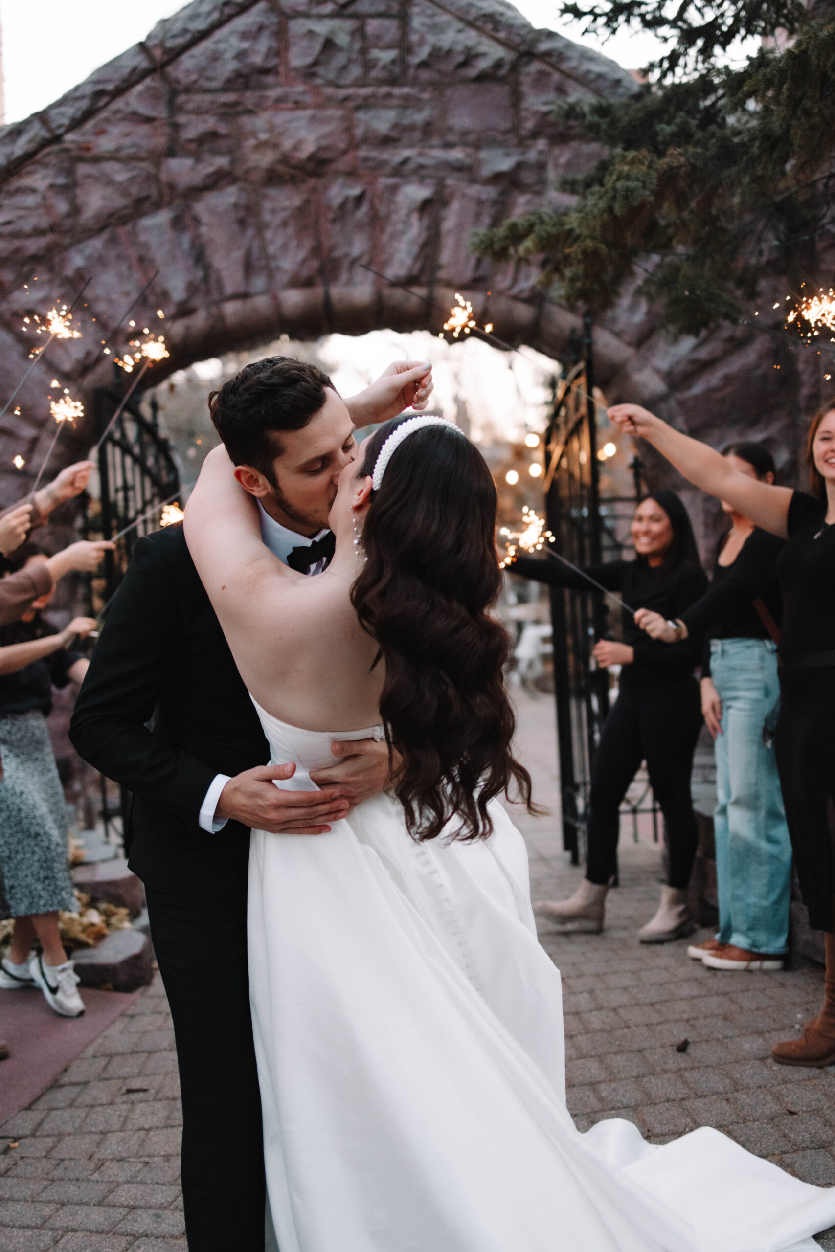 A bride and groom dip kissing below a stone archway. They are surrounded by smiling guests holding sparklers. The bride is in a white gown, and the groom is in a black tuxedo at the van dusen mansion