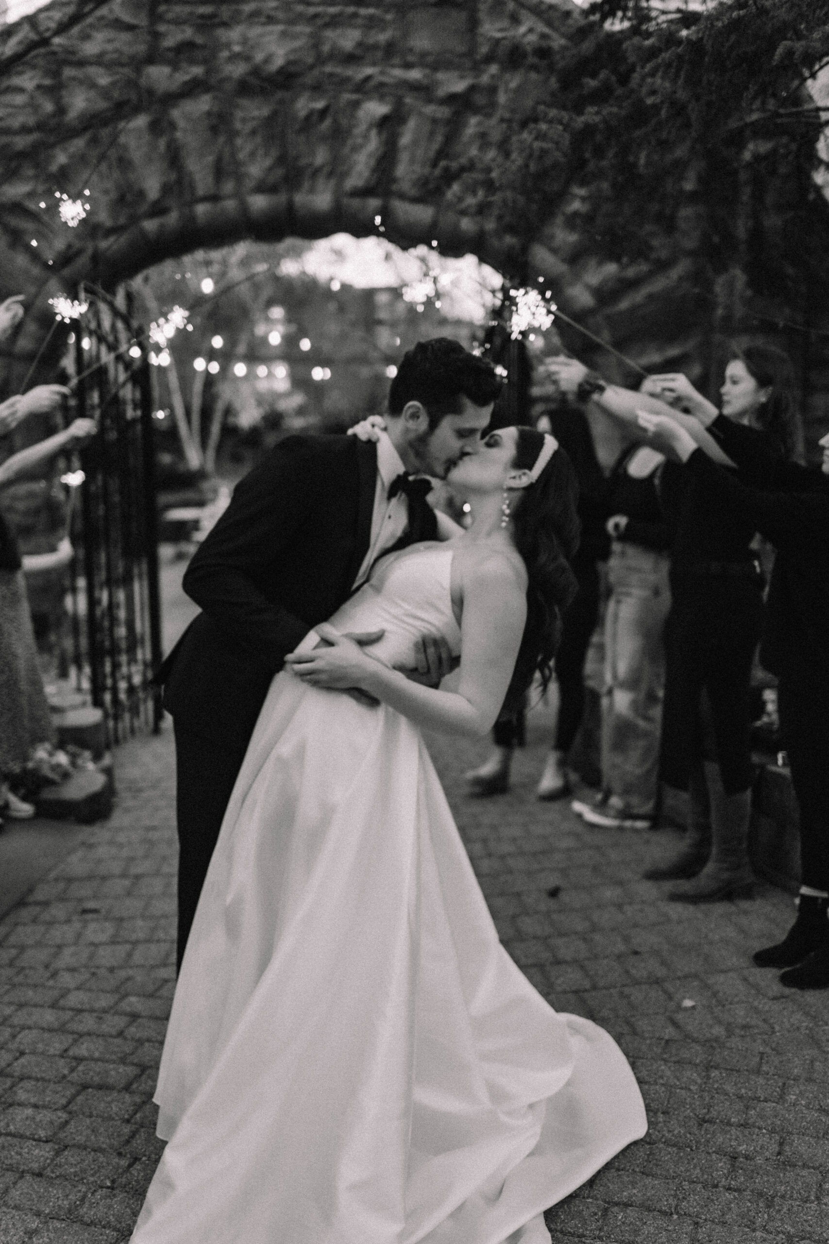 A bride and groom dip kissing below a stone archway. They are surrounded by smiling guests holding sparklers. The bride is in a white gown, and the groom is in a black tuxedo at the van dusen mansion