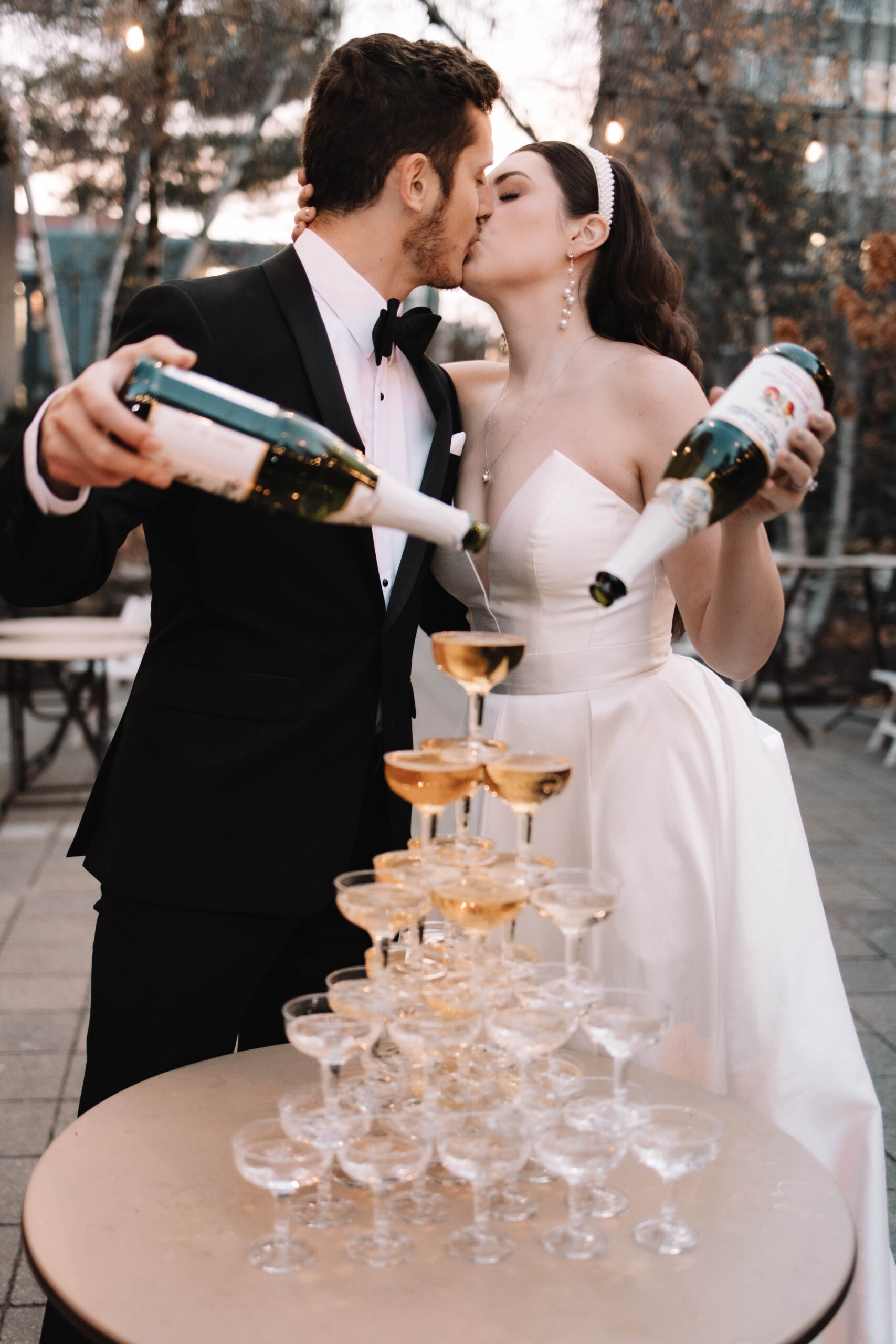 A bride and groom kiss while pouring champagne into a tower of glasses at an outdoor celebration at a wedding in Minnesota