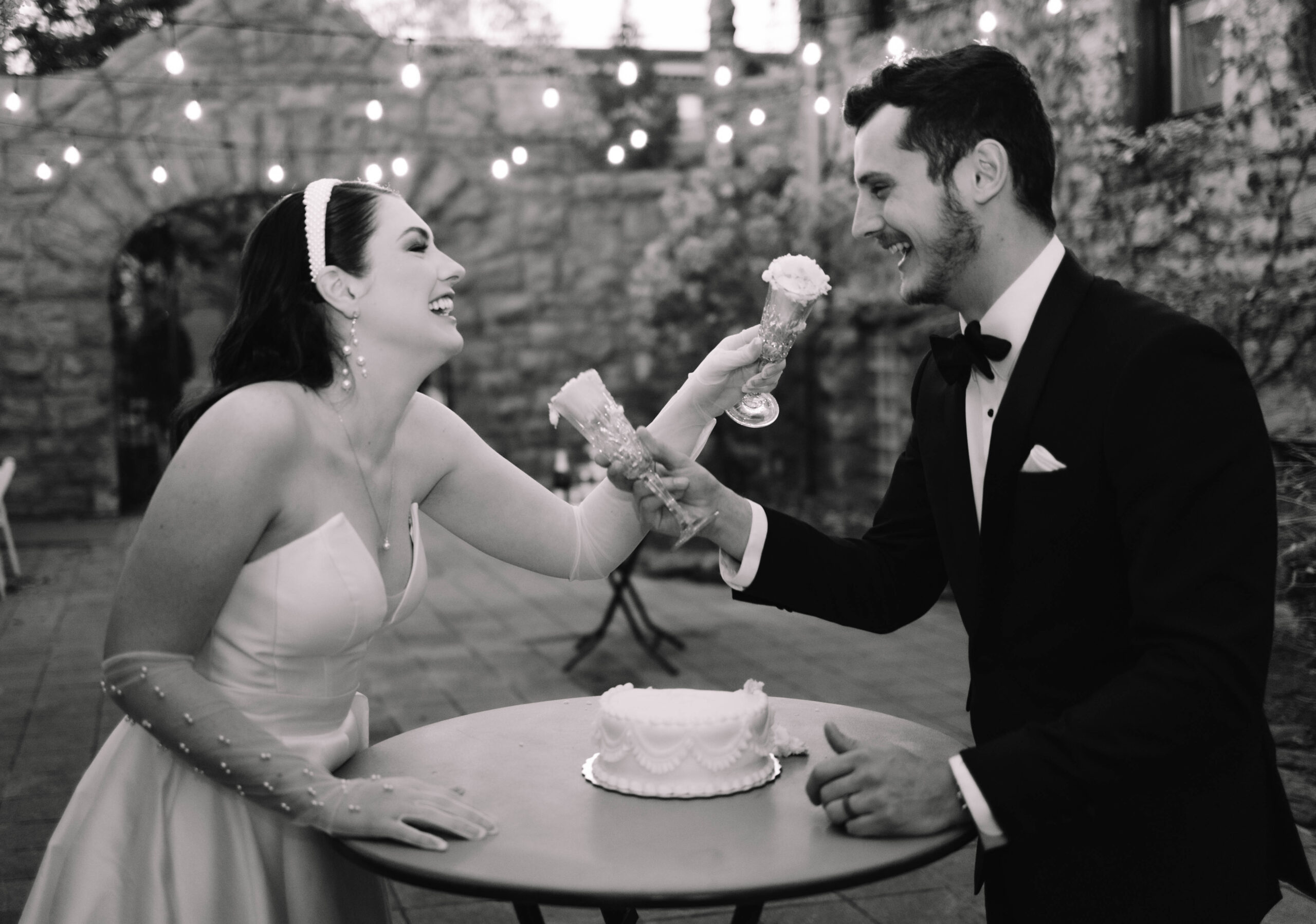 A luxury bride and groom laughing feeding each other wedding cake outside on a porch with string lights hanging in the background at the van dusen mansion