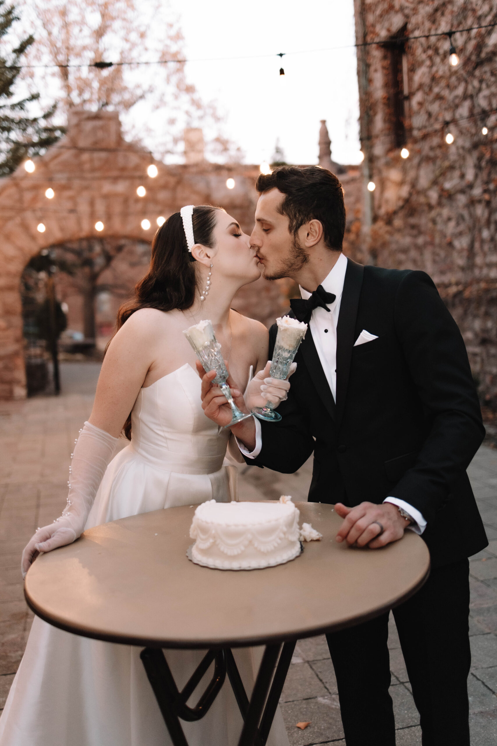 A bride and groom in luxury wedding attire kissing and standing behind their cake table that has a small white wedding cake on