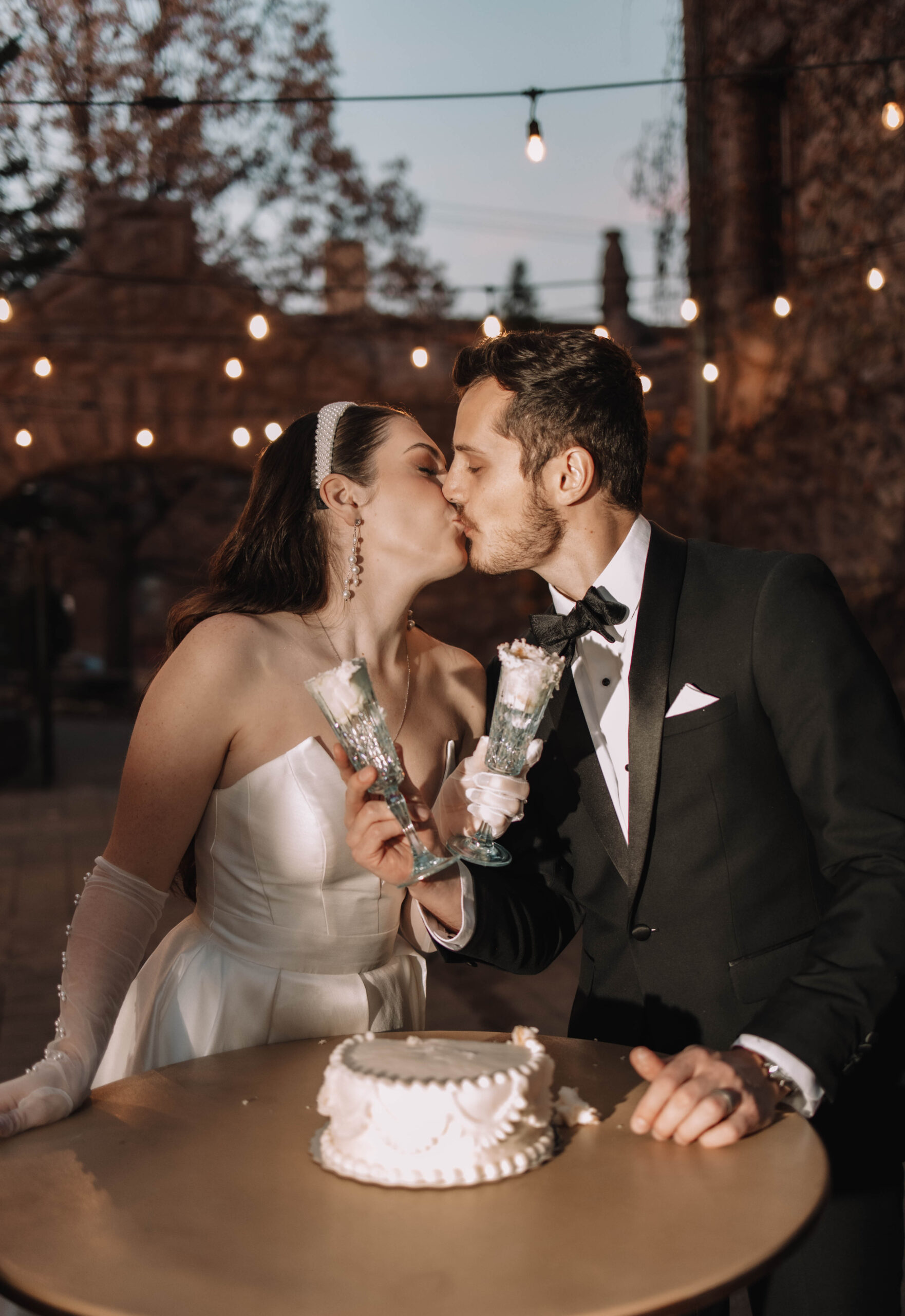 Flash photo of bride and groom kissing during their cake cutting on the back patio of a mansion. String lights are hanging behind them
