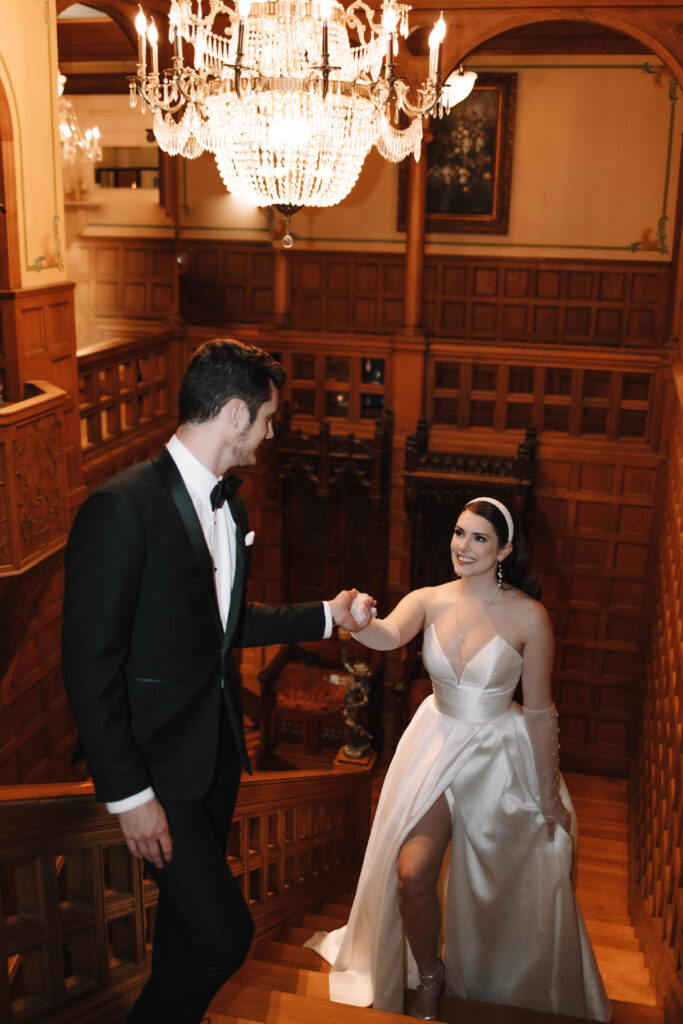 A groom leading his bride up the stairs inside an old historic mansion in Minnesota. A beautiful chandelier hangs above them.
