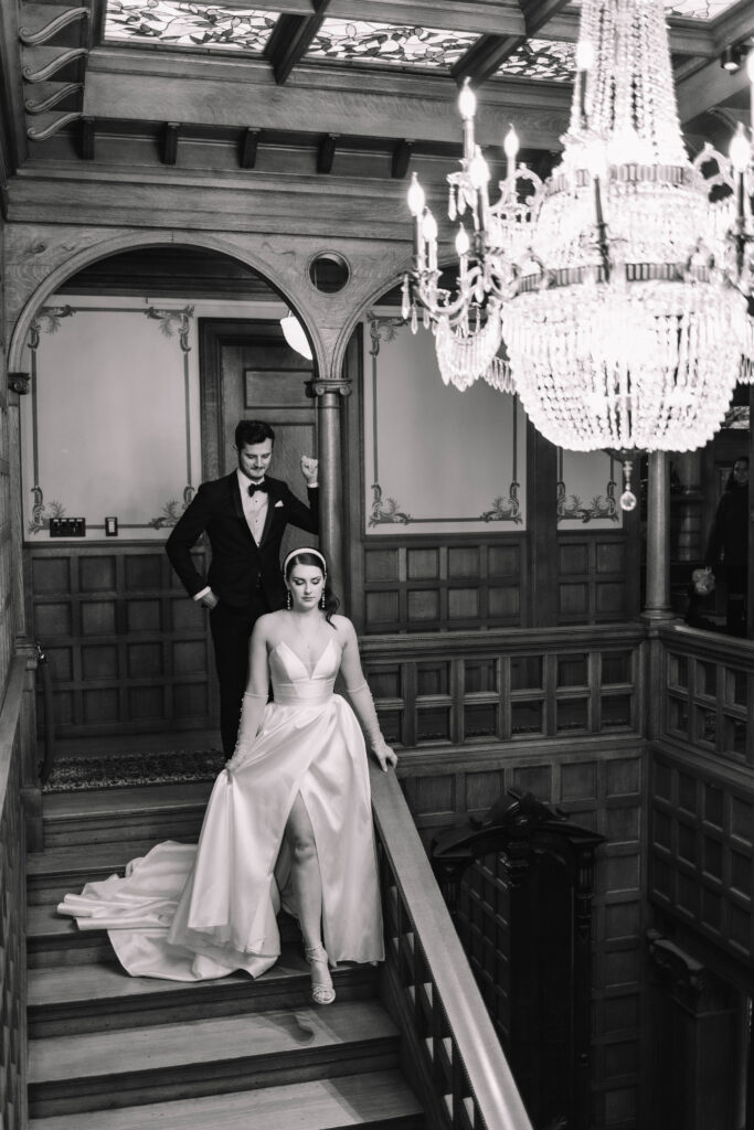 A bride and groom walking down wooden stairs in a Mansion. A chandelier hangs above them. The bride has a beautiful ballgown dress with a sweetheart neckline and a slit
