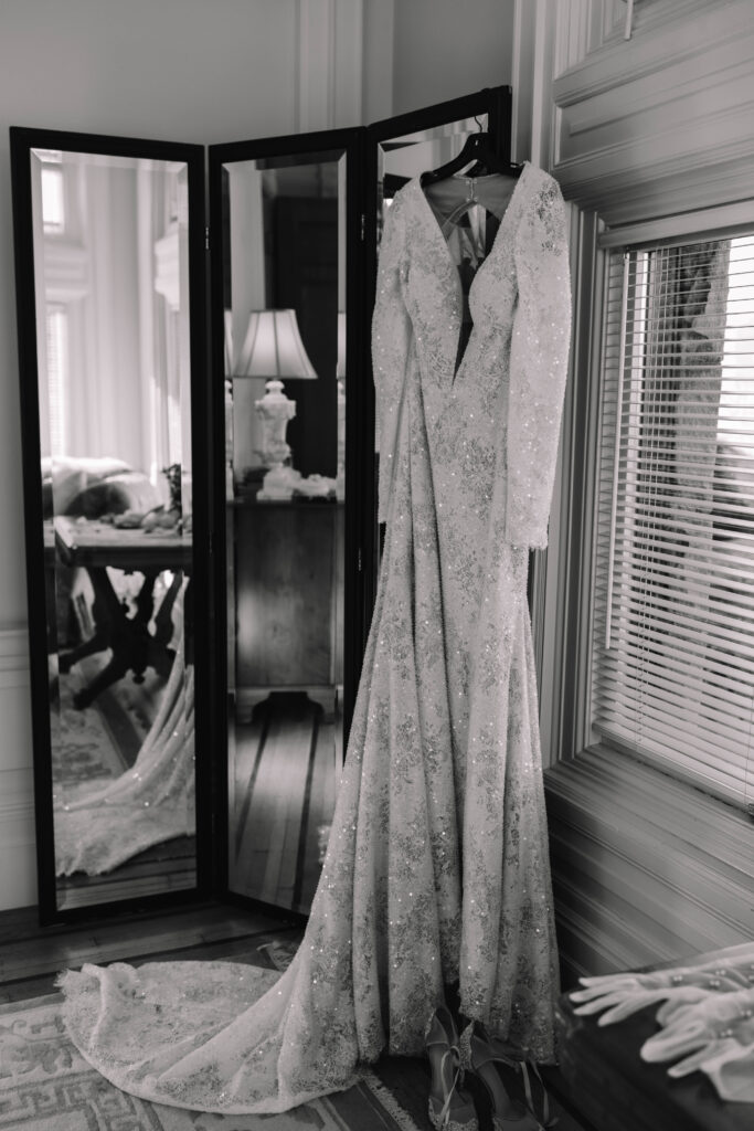 A beautiful long sleeve, open back dress with diamond embroidery hanging in front of a mirror in the bridal suite of the Van Dusen Mansion