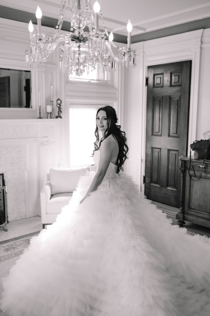 A bride in a voluptuous ballgown wedding dress standing with her back against the camera and looking over her shoulder in the bridal suite of the van dusen mansion. A beautiful chandelier hangs above her