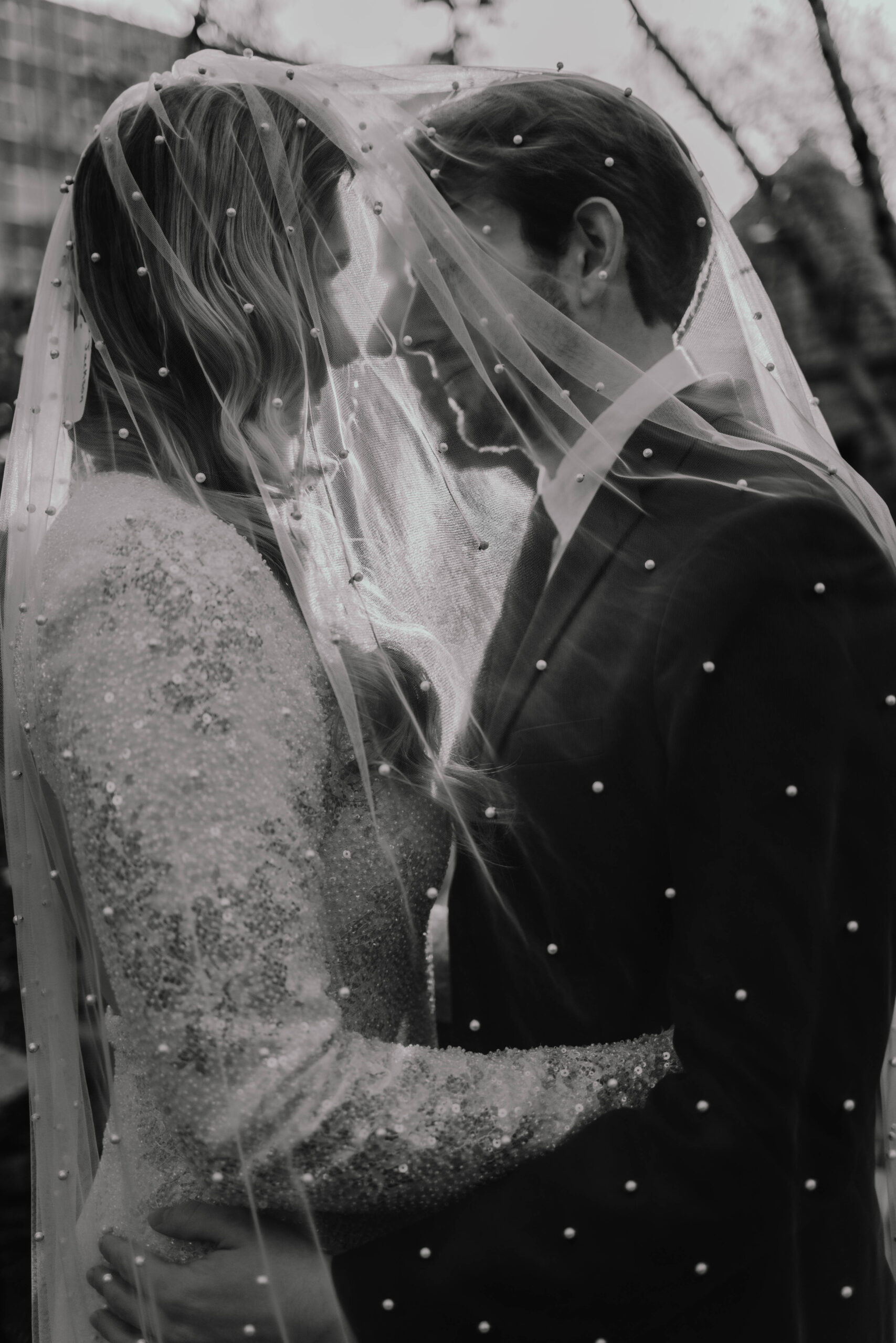 A bride and groom kissing underneath a pearl embedded veil as the sun sets behind them creating a silhouette