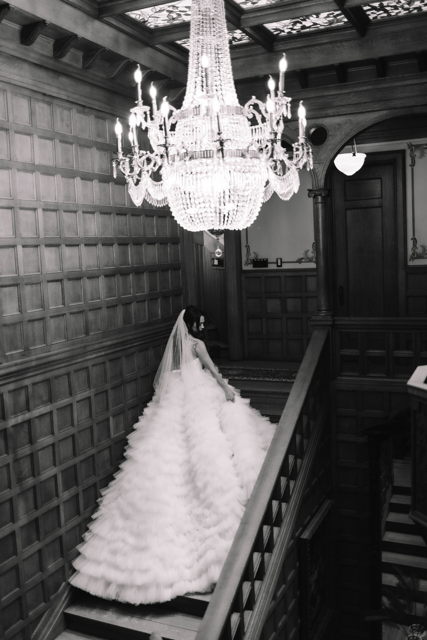 A bride in a voluptuous wedding dress walking up the stairs inside a mansion with a large chandelier hanging above her
