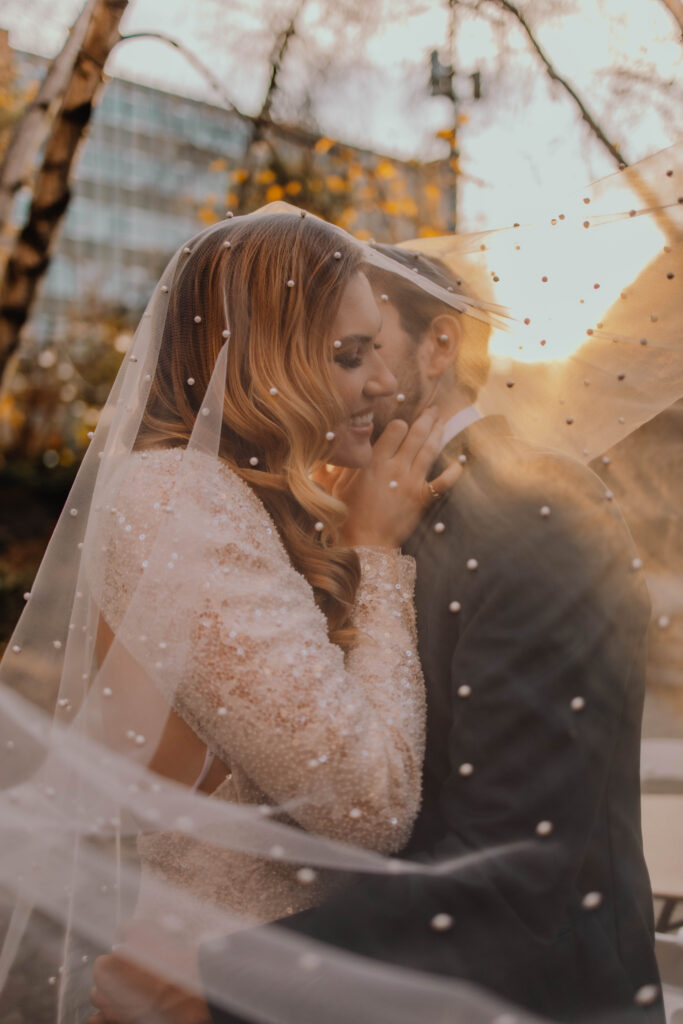 Groom holding his bride and kissing her on the cheek while they're both underneath a veil covered in pearls as the sun shines behind them