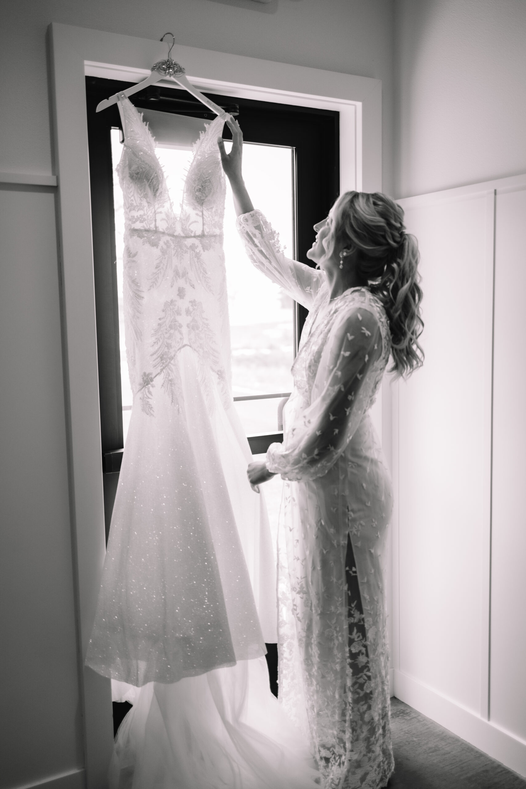 Wedding shot of a Bride admiring her wedding dress hanging from a door next to a window, with the light shining through illuminating the details of the dress