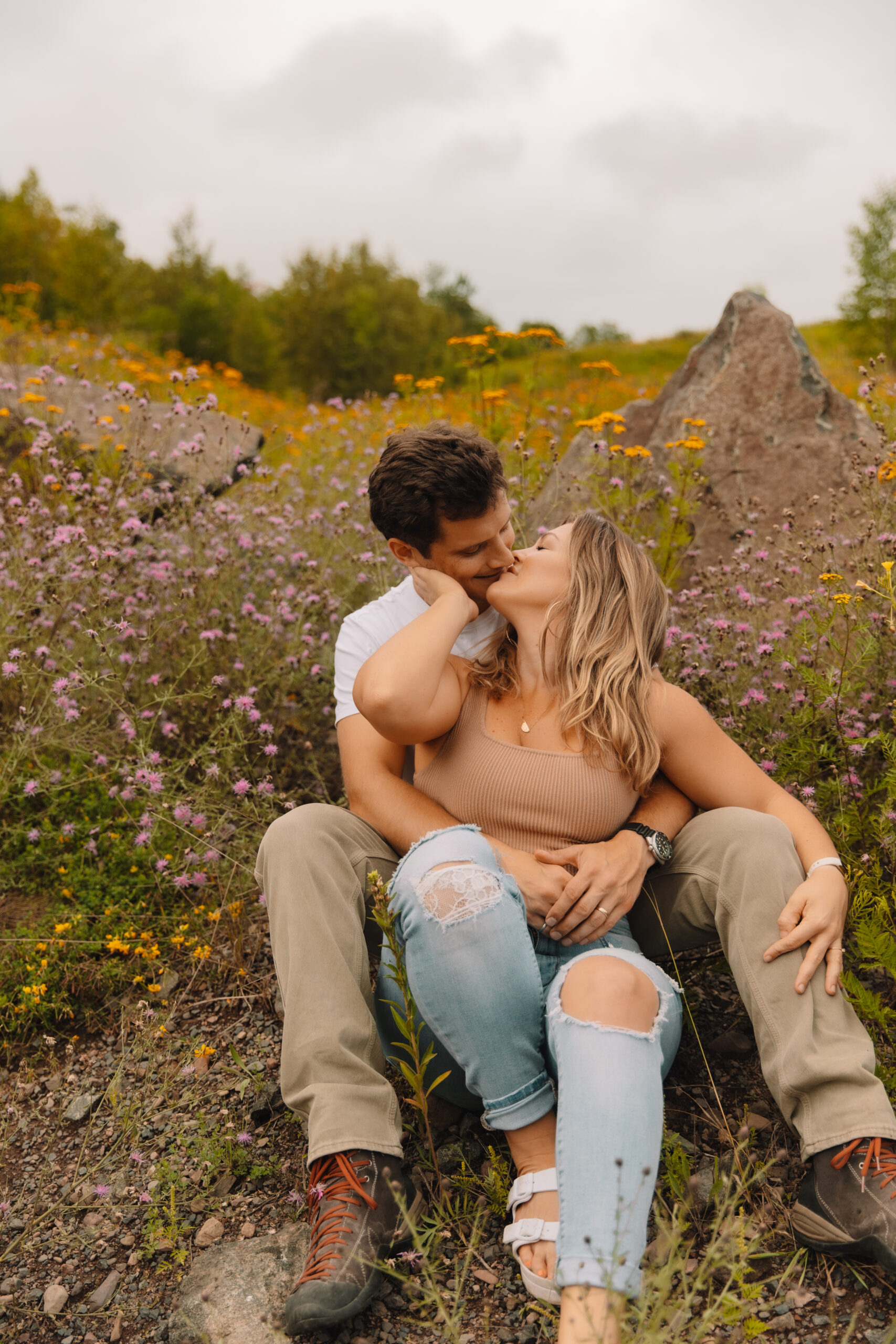 A couple sits among wildflowers and rocks, with the man embracing the woman from behind as they kiss.