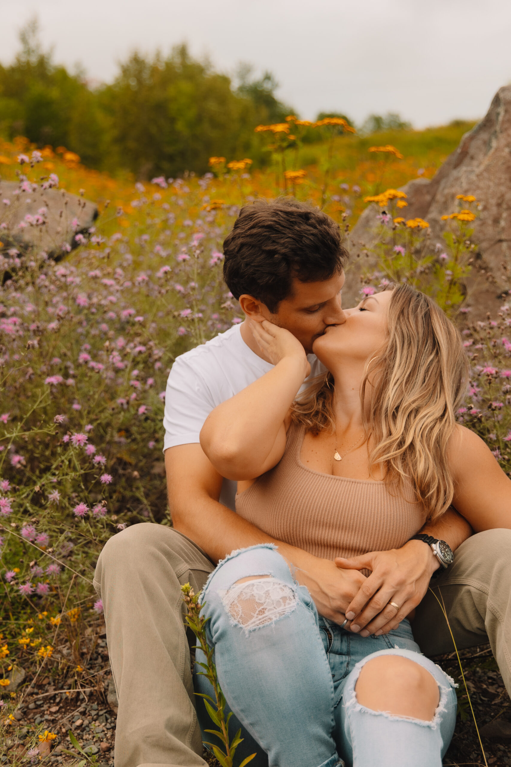 A couple sits among wildflowers and rocks, with the man embracing the woman from behind as they kiss.