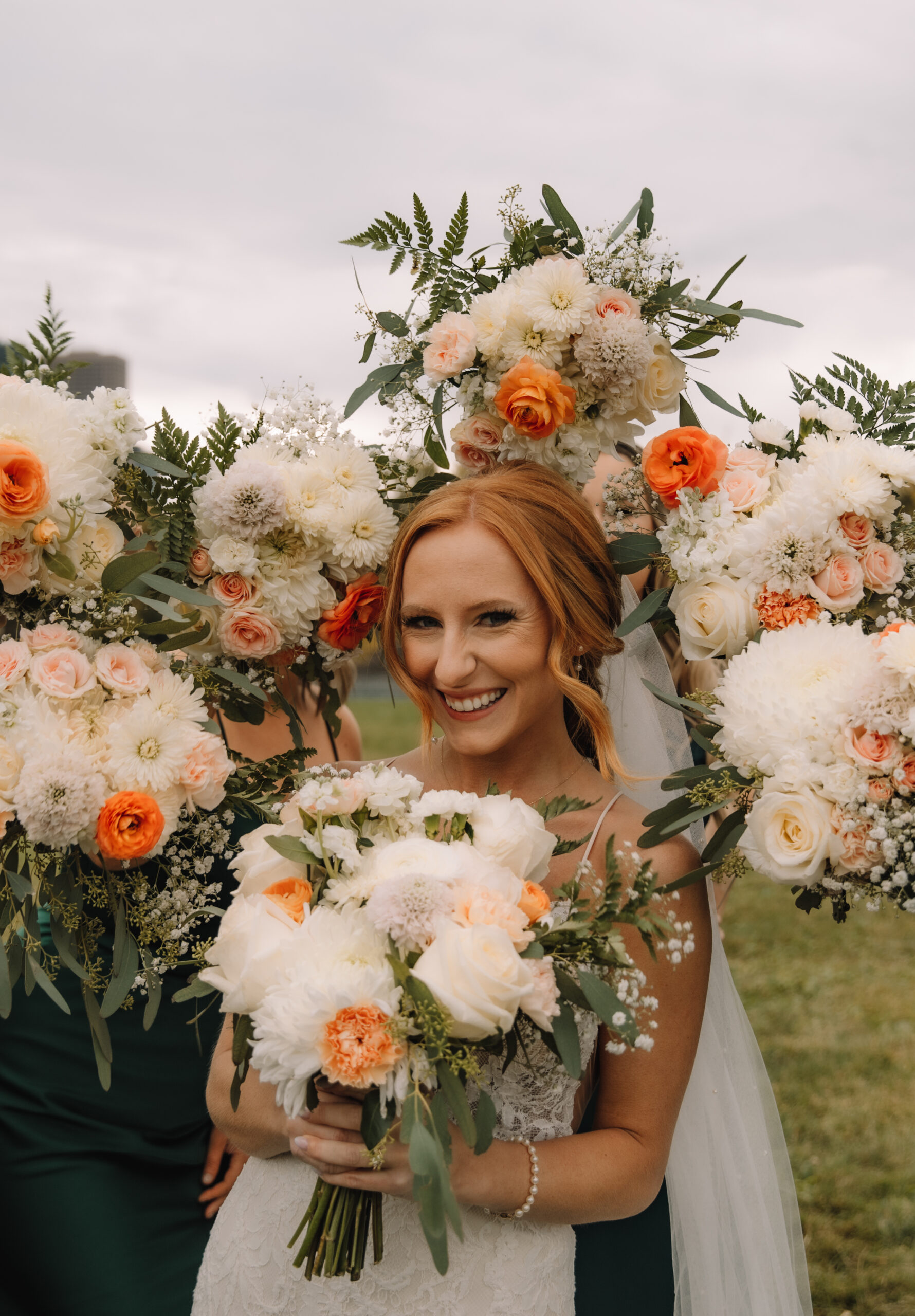 Bride smiling, holding a bouquet of white and peach flowers, with bridesmaids in teal dresses carrying similar bouquets, outdoor setting.