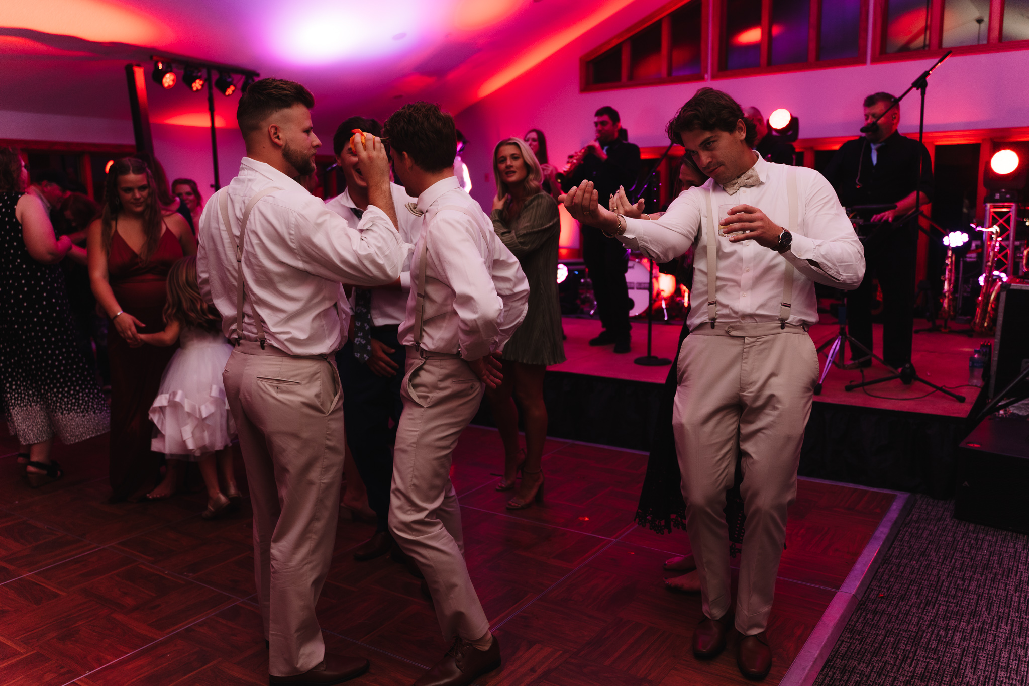 Four men in semi-formal attire dancing energetically with each other on a dance floor at a lively evening event, with other guests and a live band in the background at a wedding in Minnesota