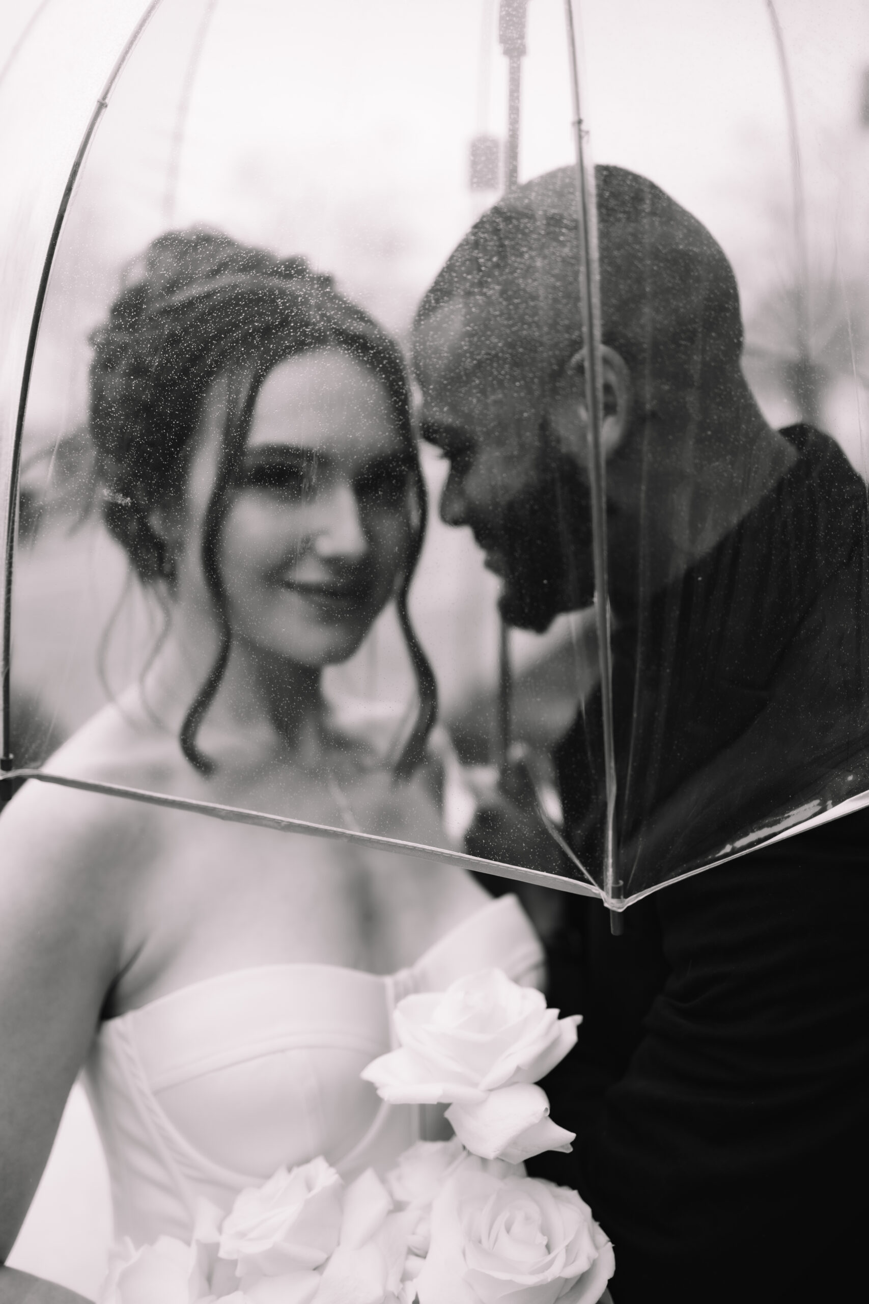 A bride and groom standing underneath a clear umbrella covered with rain droplets 