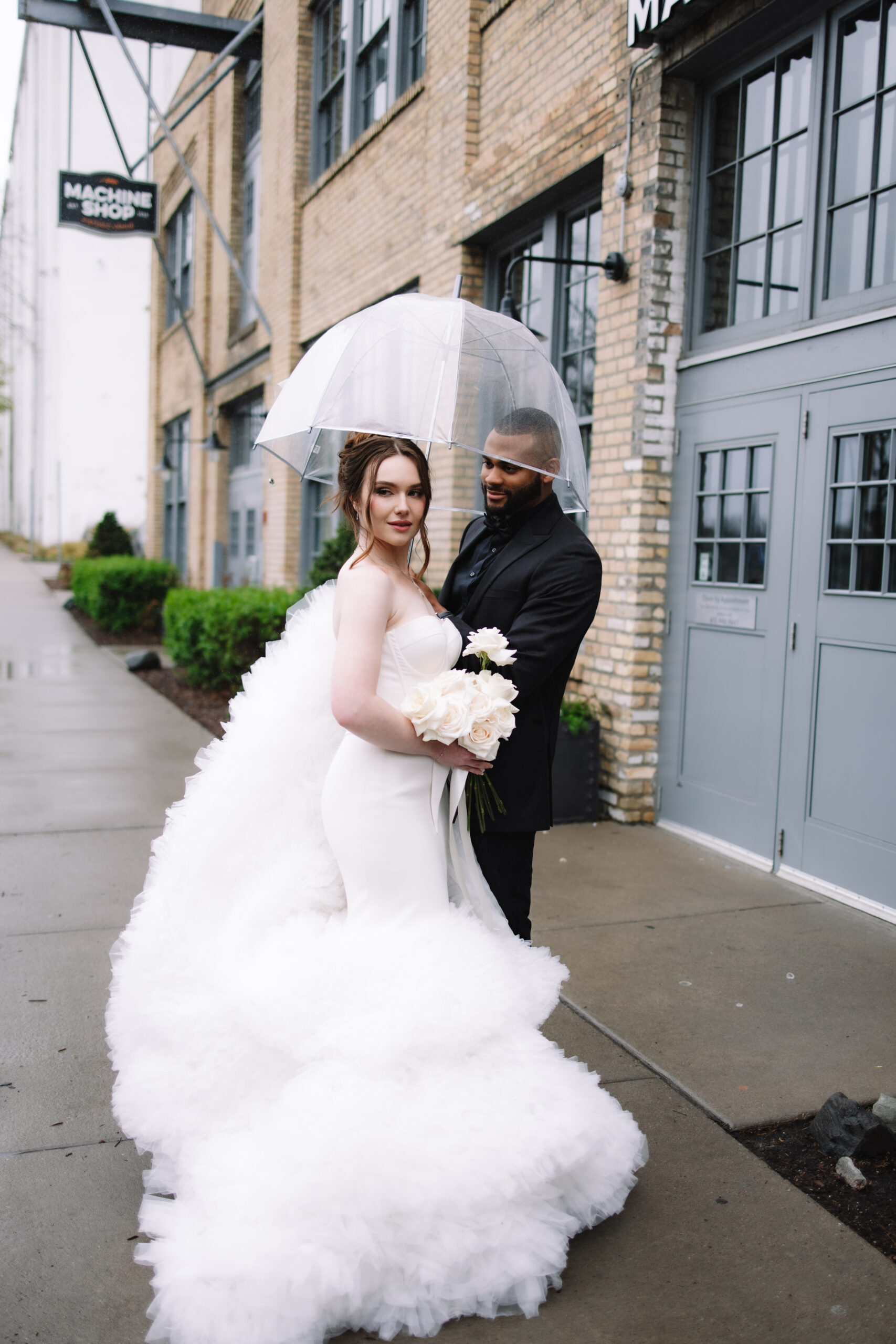 A bride in a voluptuous wedding dress standing with her groom under an umbrella in front of The Machine Shop wedding venue in Minneapolis
