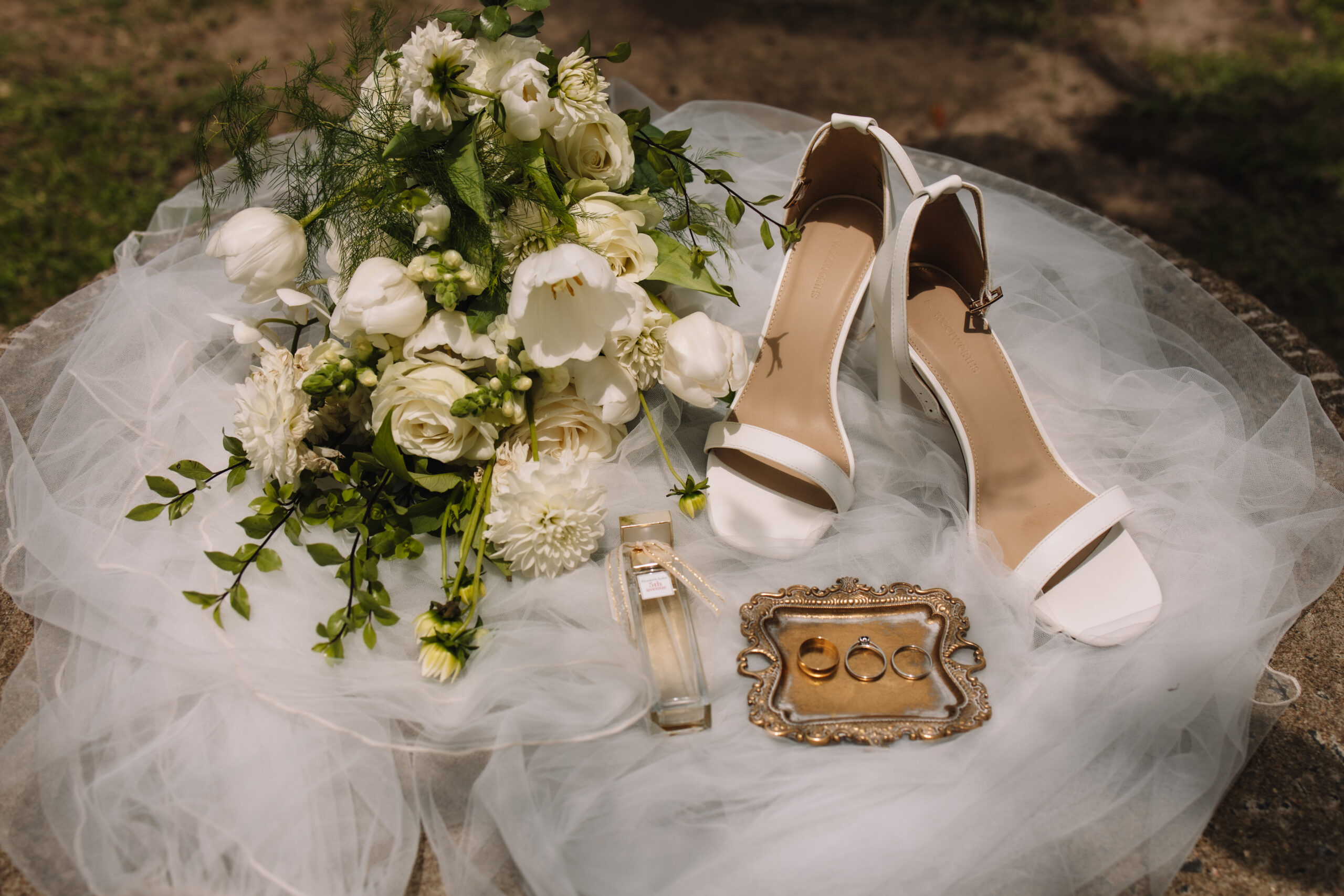Elegant, modern wedding flat lay details in the sun. The flat lay details includes cheek wedding shoes, a white and green bouquet, perfume, a veil and rings in a vintage ring holder