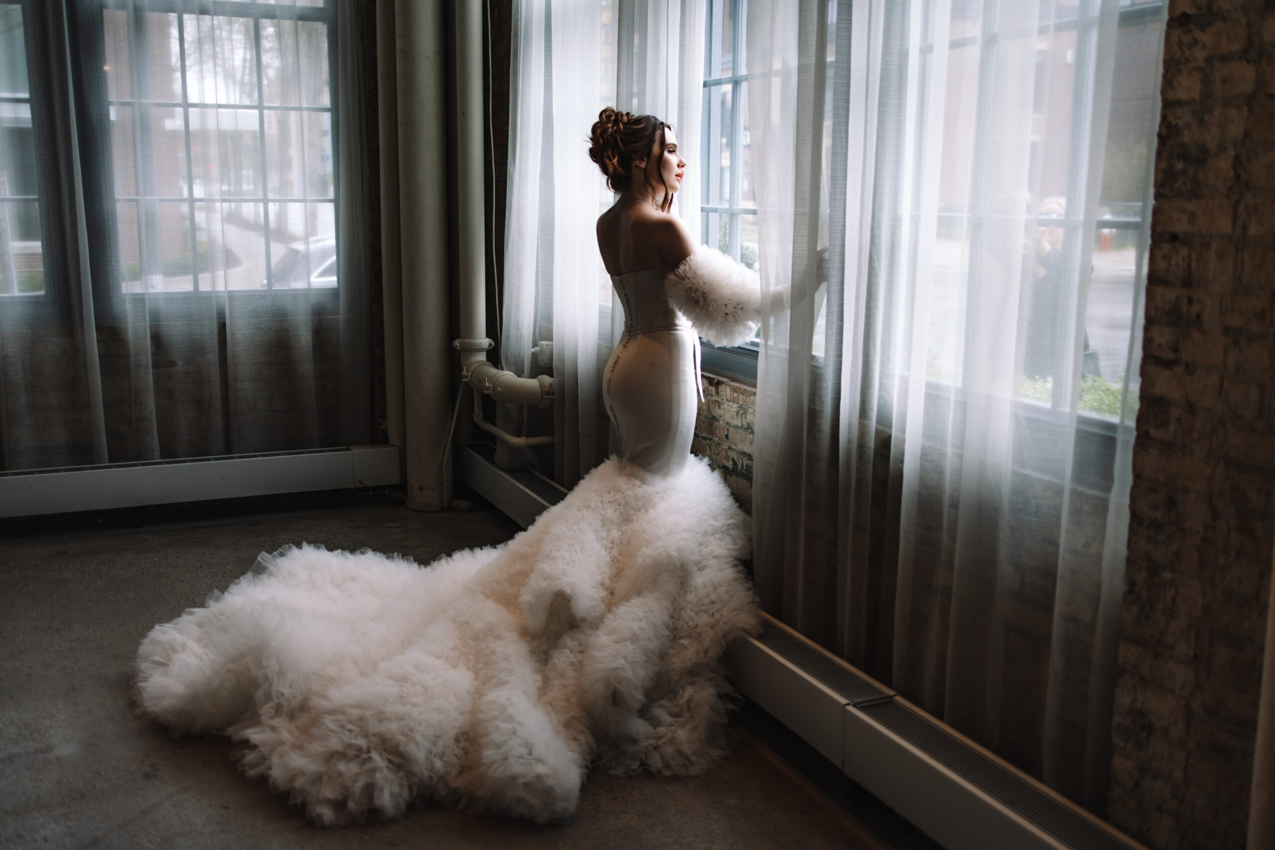 A modern, elegant bride in a voluptuous dress with sleeves, staring out the window as the light illuminates her face