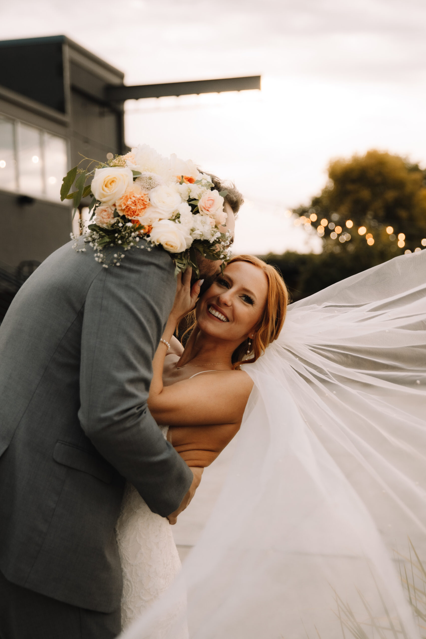 Bride smiling as her husband kisses her neck. Her bouquet with white and peach flowers leans over his back