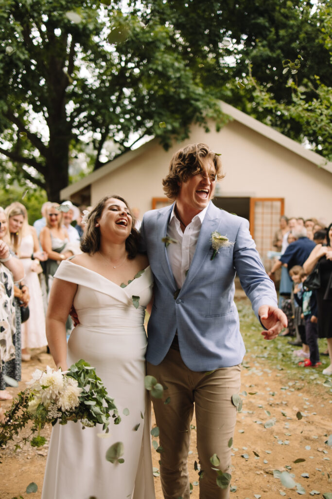 Bride and groom walking back down the aisle laughing while guest throw eucalyptus confetti petals at them
