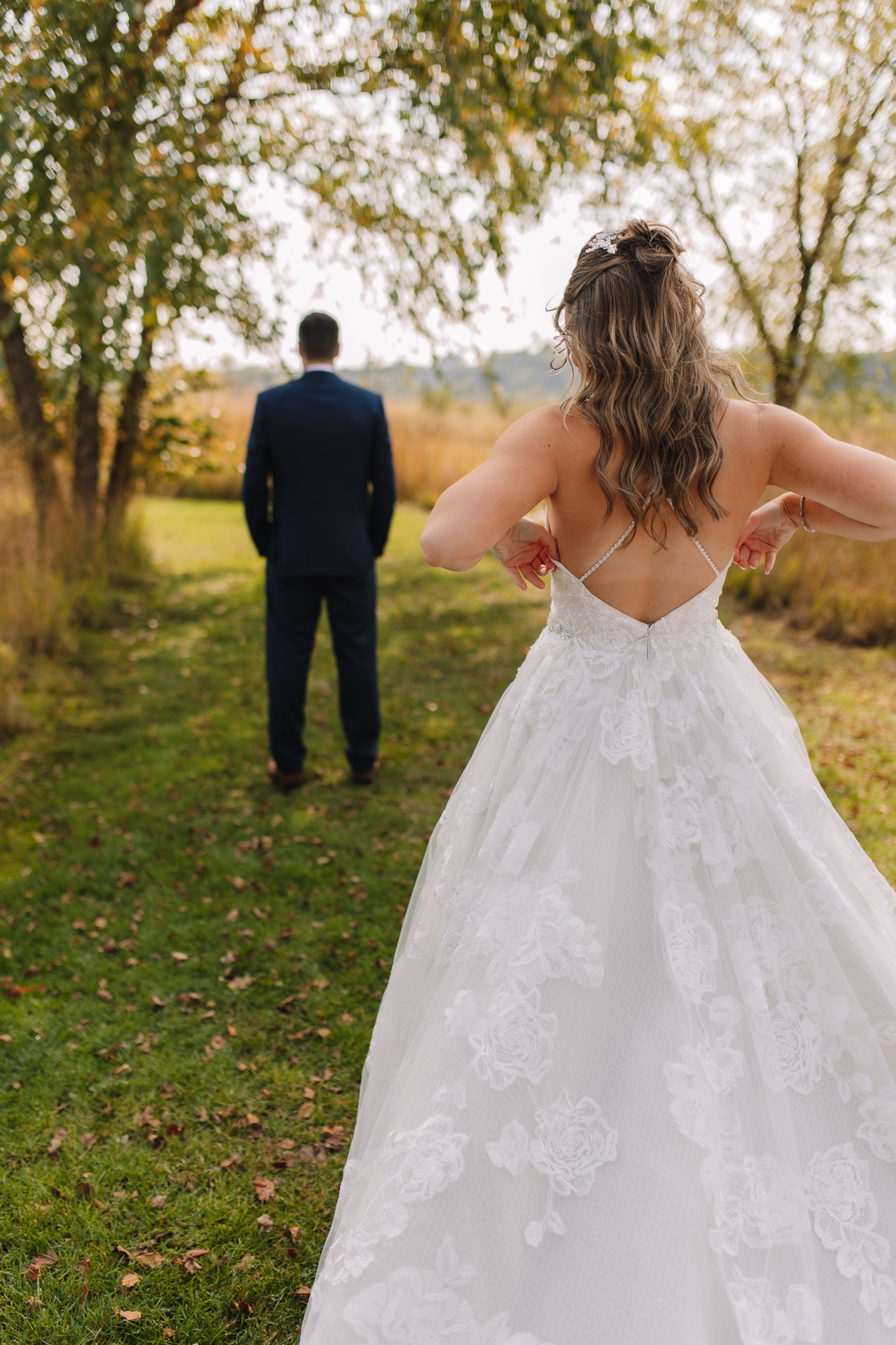 Bride in an open back wedding dress with criss cross straps standing behind her groom while he faces away waiting to turn around and see his bride for the first time in her wedding dress