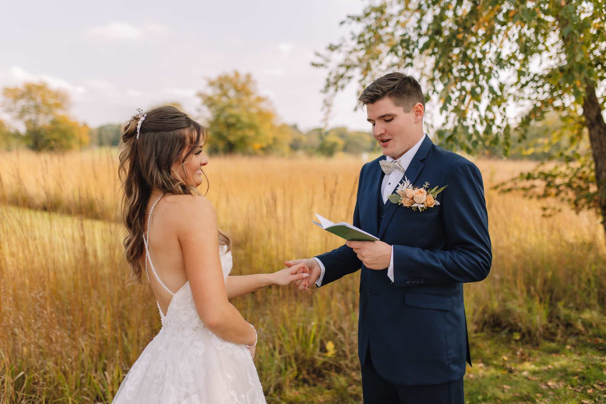 Groom holding his Bride's hand as he reads her private vows from his vow book in a field