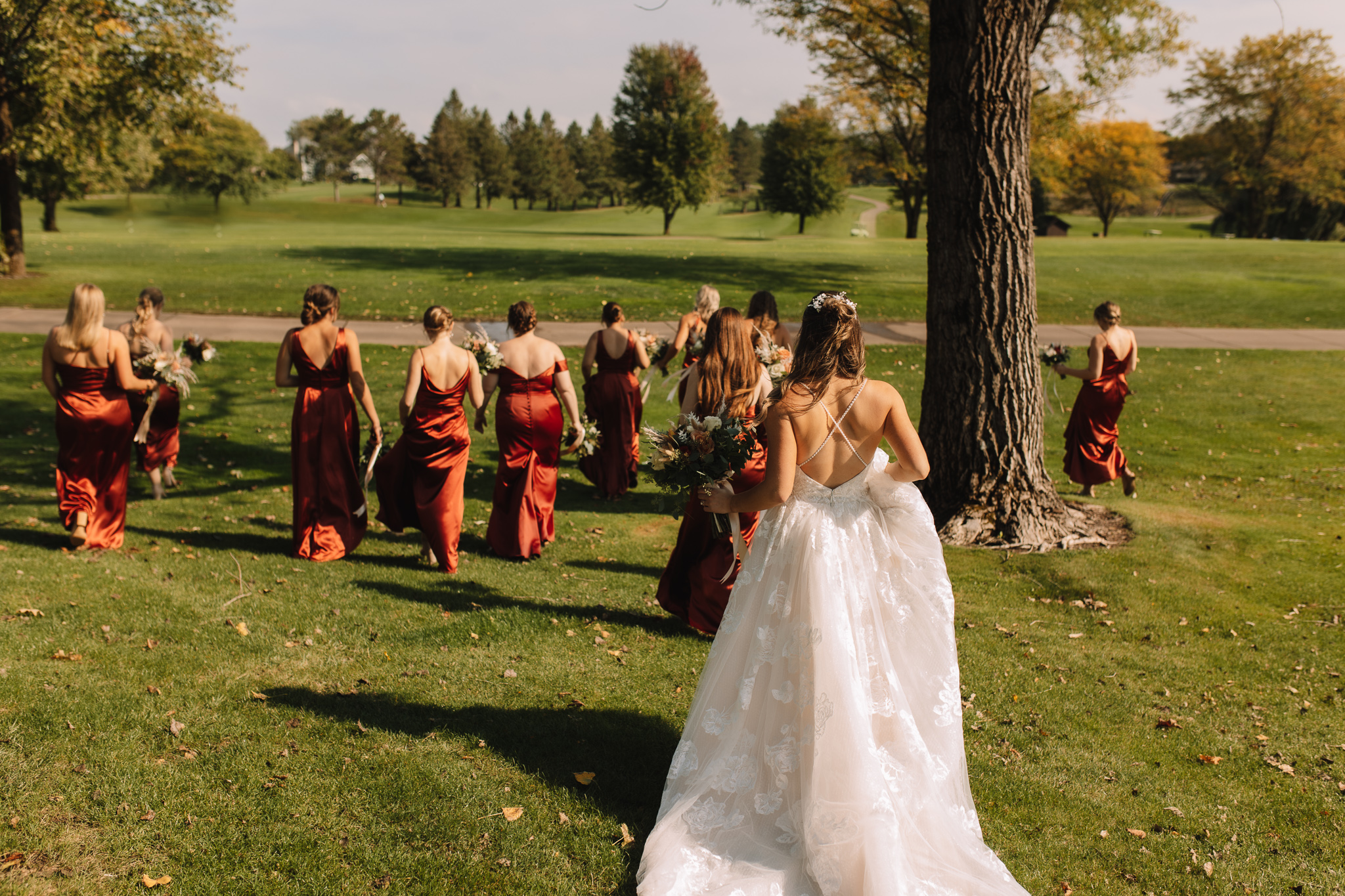 A bride in an open back wedding dress with thin criss cross straps walking  on a golf course with all her bridesmaids
