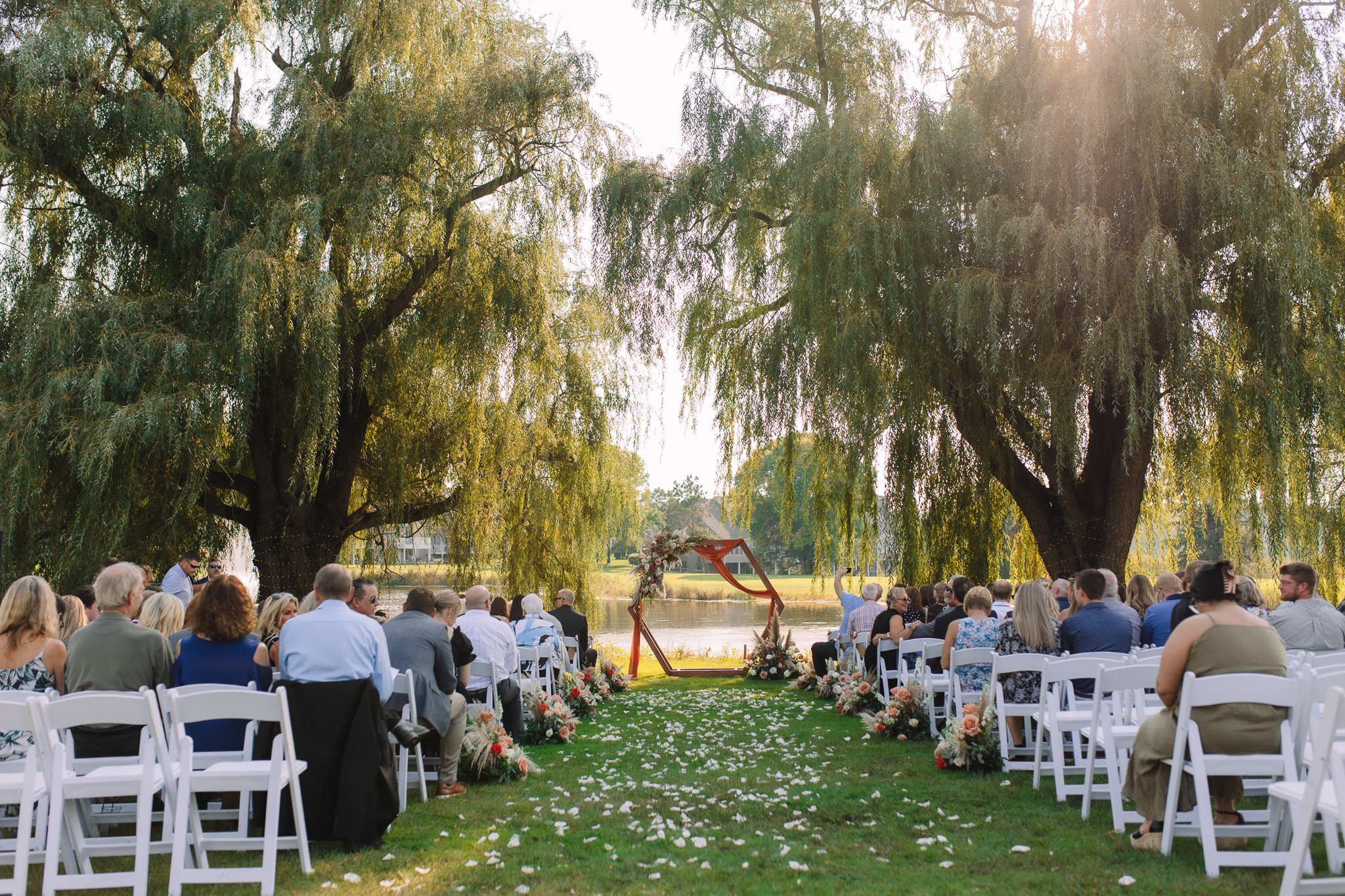 Stunning outdoor ceremony set up between willow trees. The sun shines through the trees and the pond glisters in the background