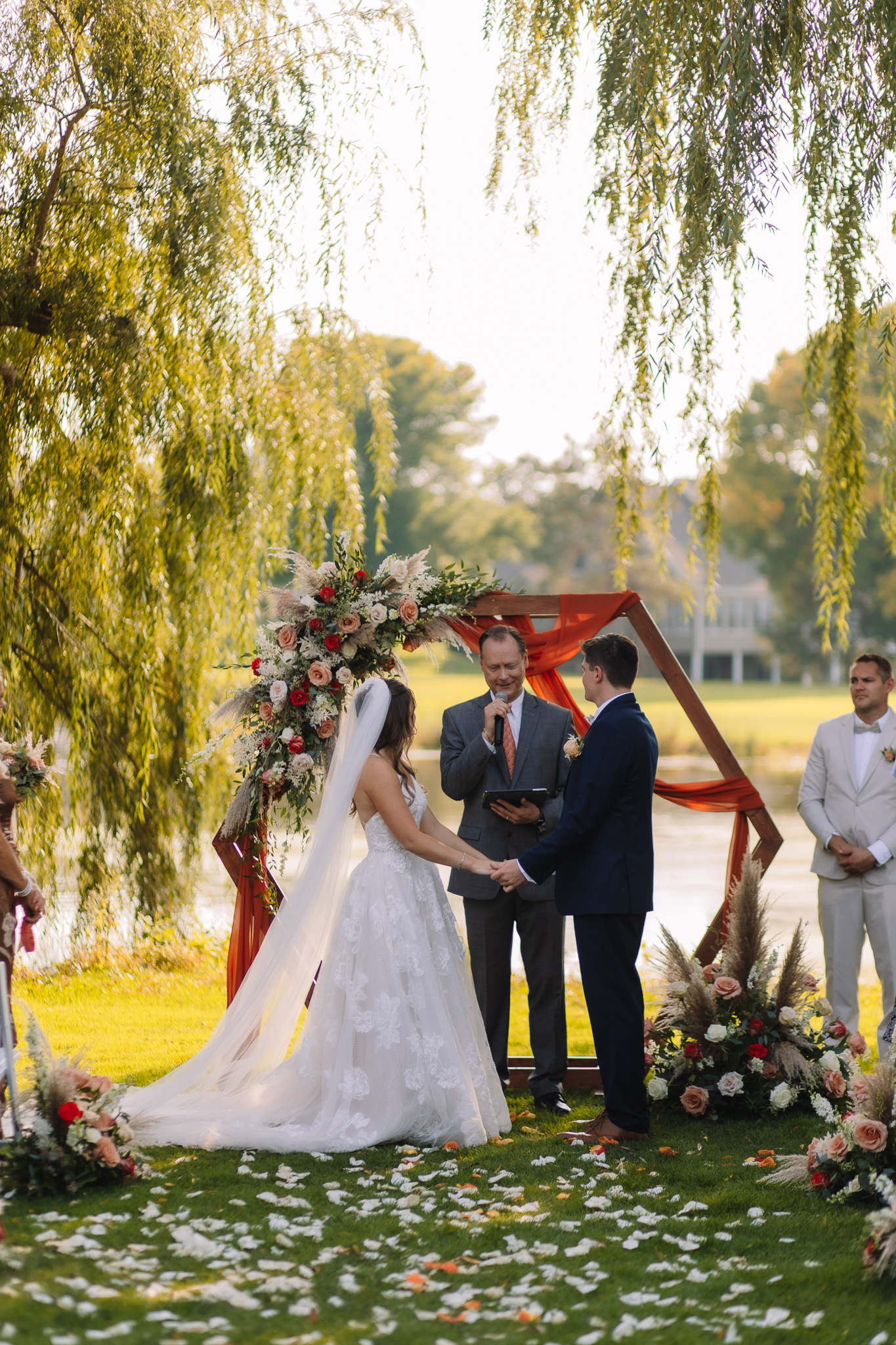 A bride and groom holding hands in front of a hexagon wooden arch. Two willow trees hang beside them and the aisle is covered in rose petals