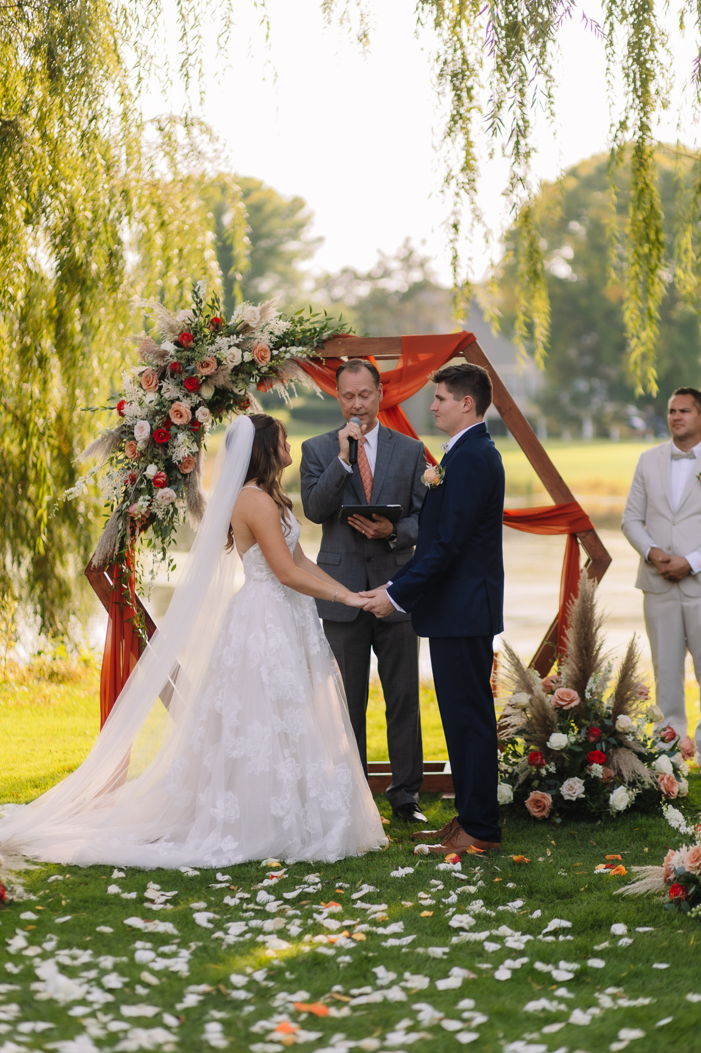 A bride and groom standing together in front of the alter holding hands. They are surrounded by willow trees and lush green grass. Flower petals are scattered down the alter of their outdoor wedding ceremony in Stillwater