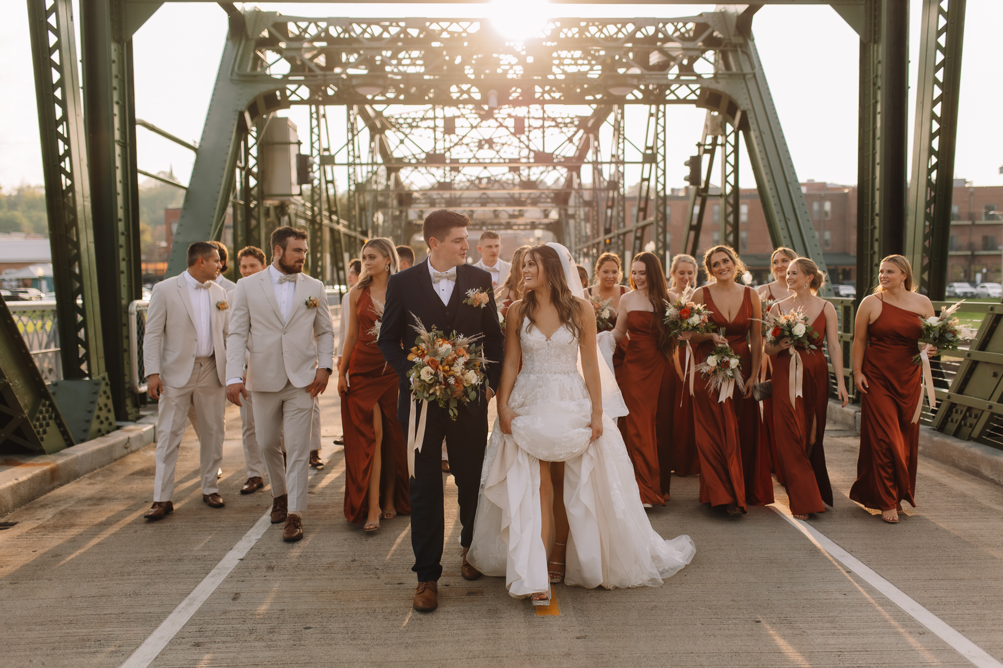 A bride and groom walking side by side under the Stillwater bridge in Minnesota with the bridal party following behind as the sun sets