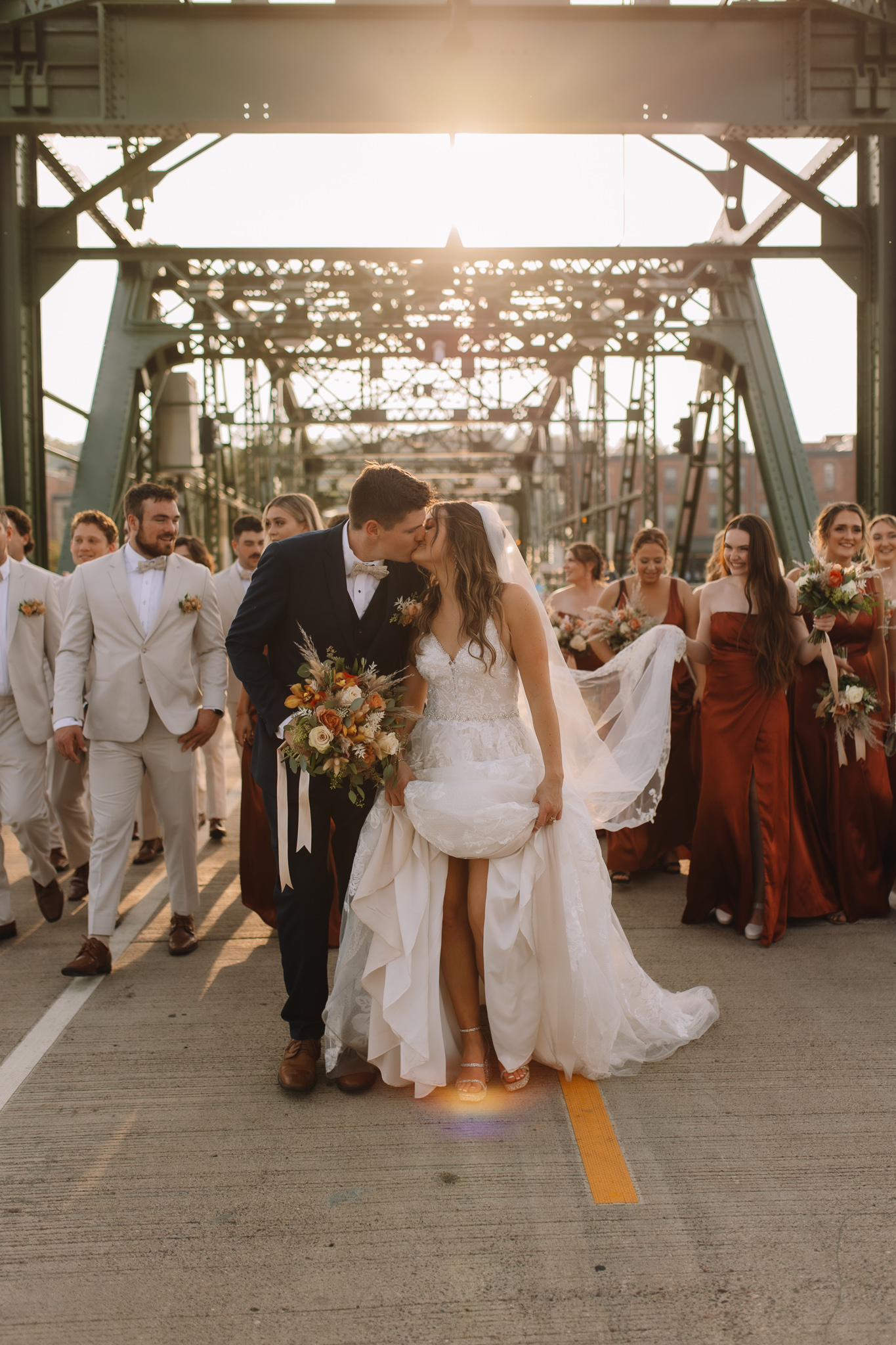 Bride and groom kissing during golden hour on the Stillwater Bridge crossing the Mississippi river with their wedding party following behind