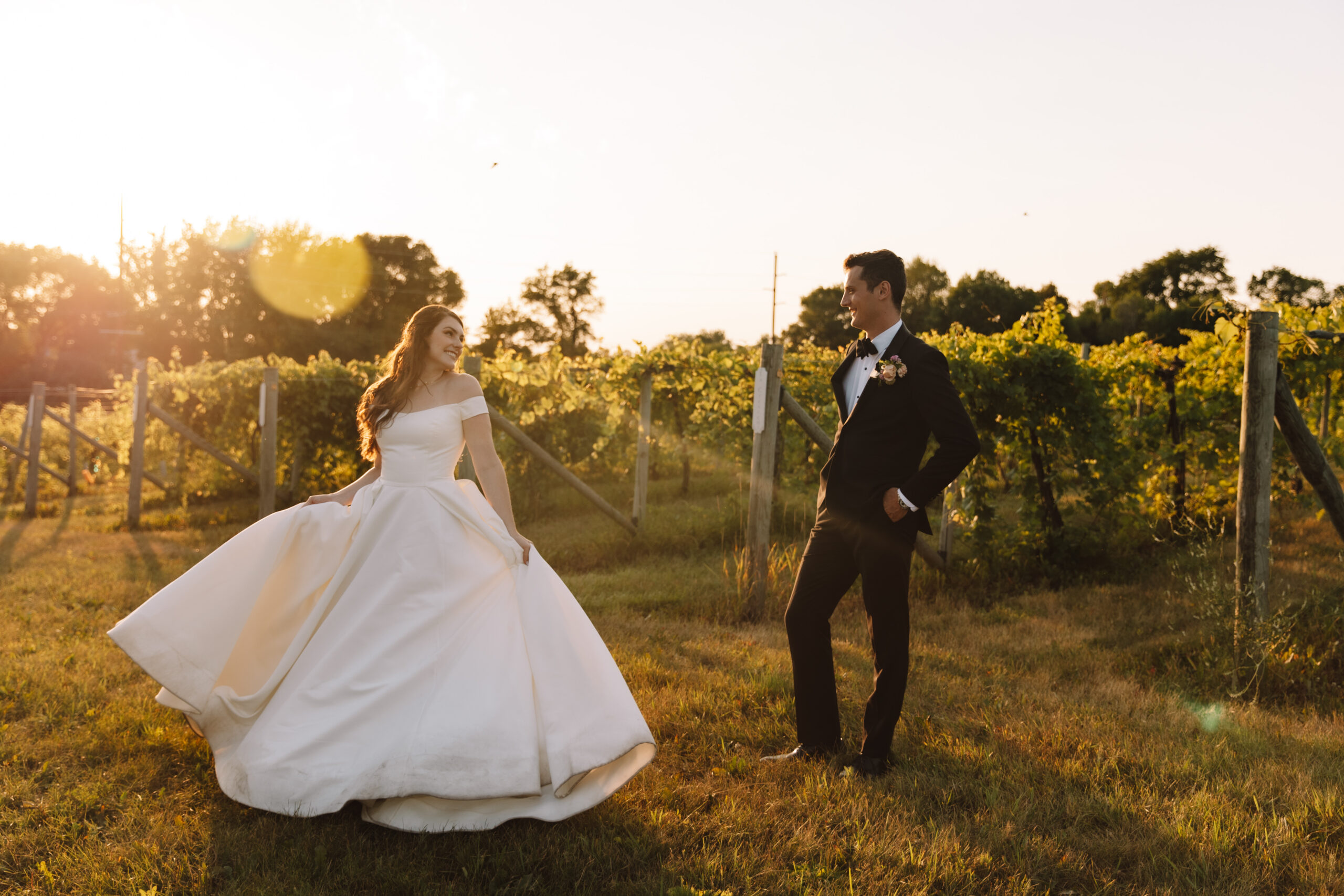 A beautiful bride twirling in her wedding dress in front of her husband in between the vineyards as the sun sets.