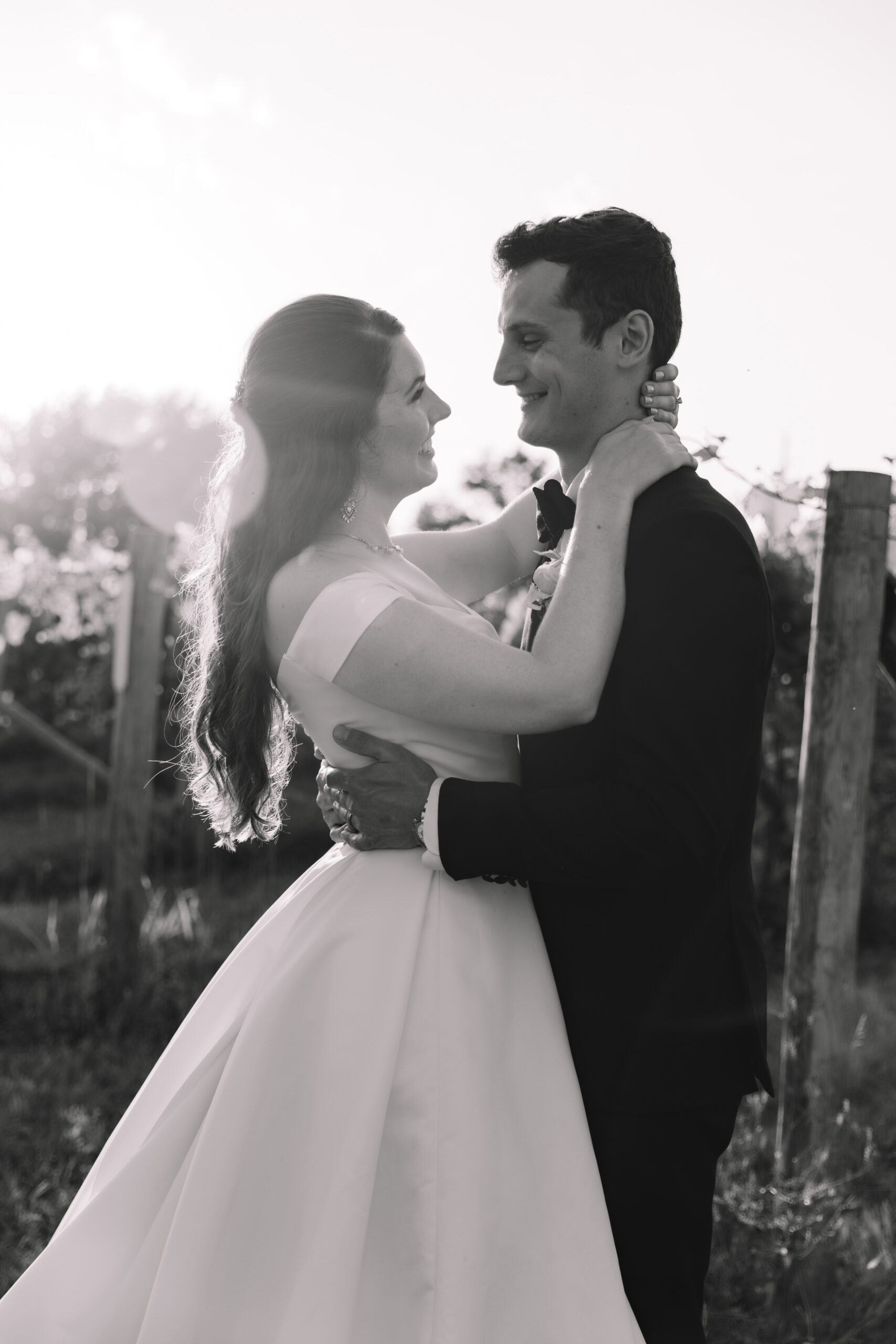 A black and white photo of a bride and groom lovingly embracing in a vineyard at sunset, with soft sunlight filtering through in the background at a wedding in Minnesota