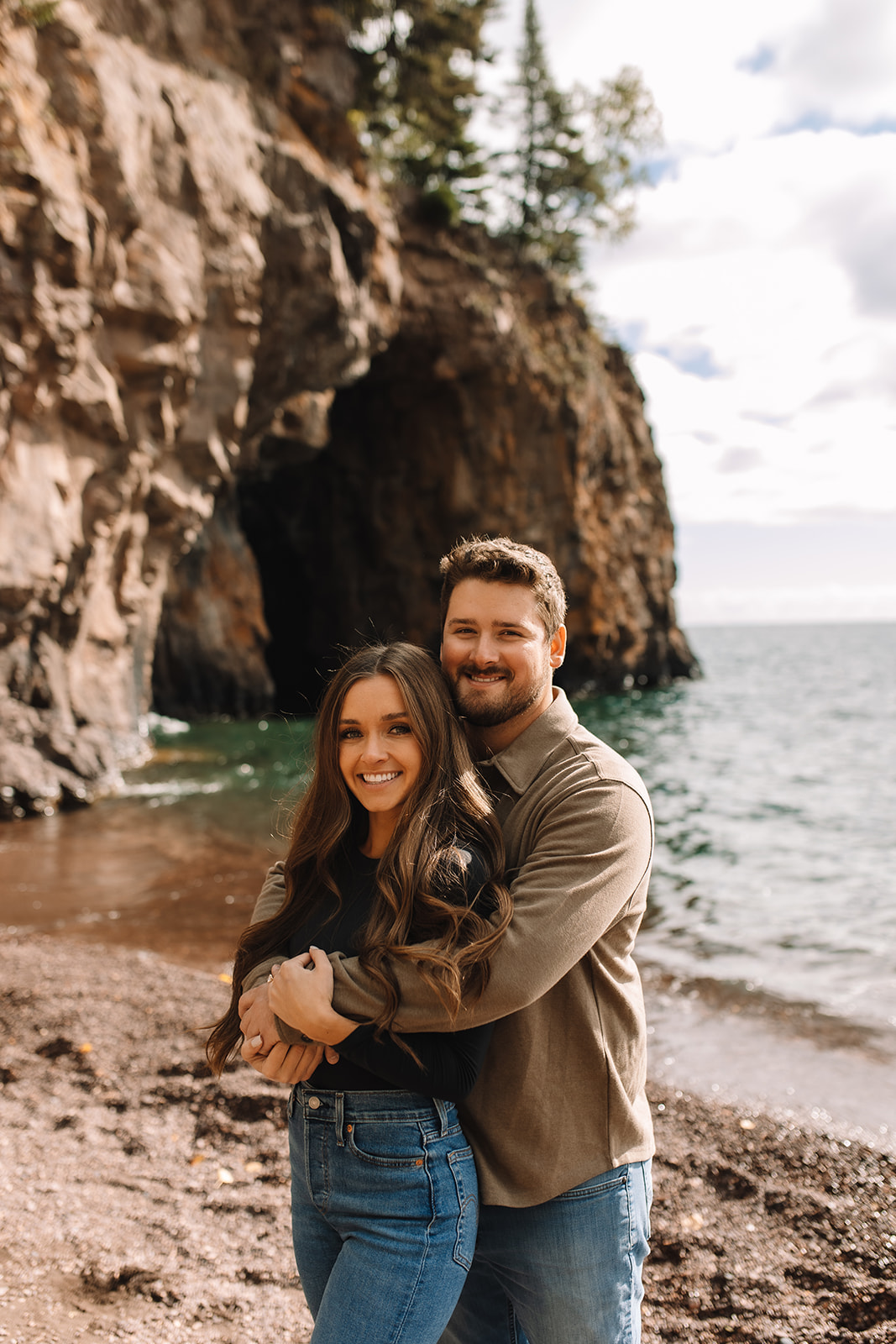 A guy holding his girl from behind while they both smile at the camera for their engagement photos on the North Shore in Minnesota