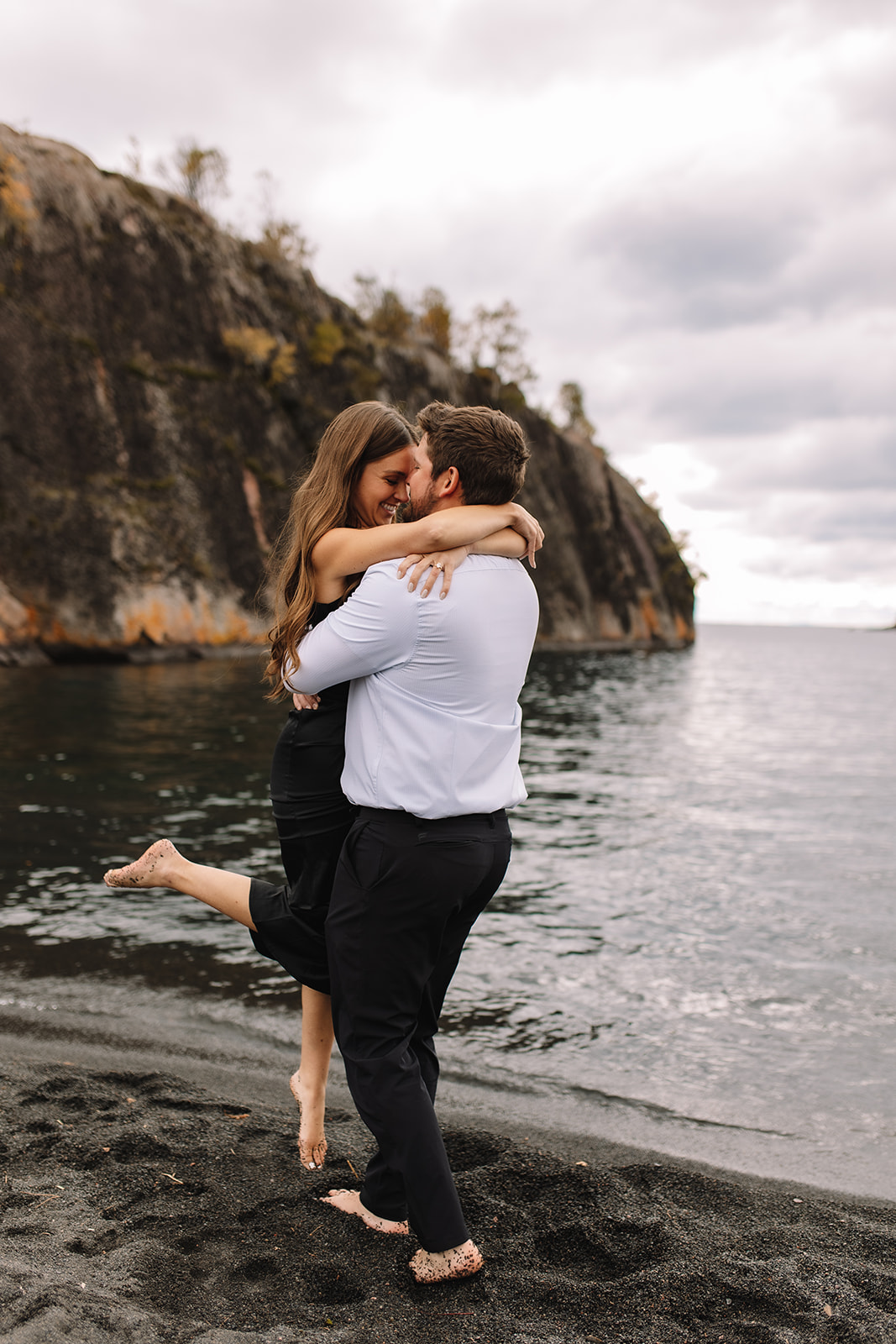 A guy picking up his girl with her arms around his neck and her leg kicked up, while they spin around on the black sand beach in Minnesota