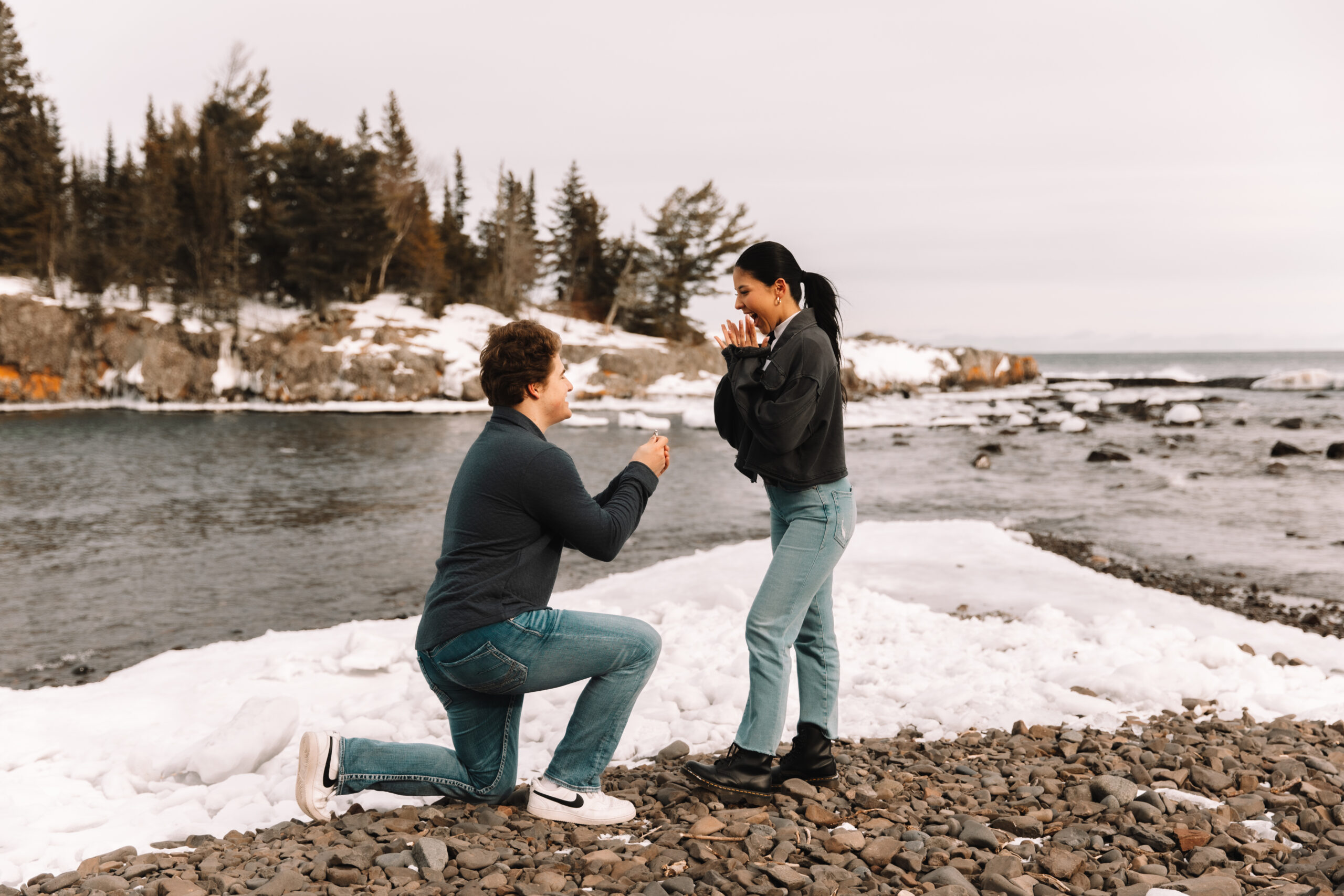 A guy proposing to his girl on a pebbled black sand beach with a beautiful lake in the background surrounded by snow.