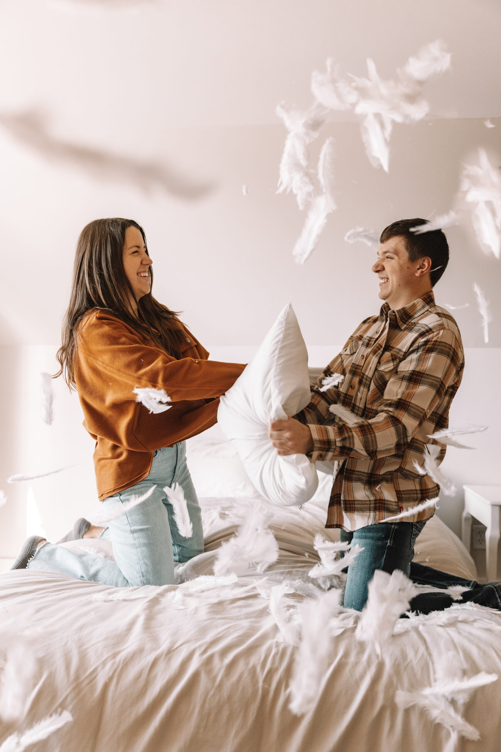 Newly engaged couple pillow fighting on a bed in long sleeve button ups and jeans for their winter engagement