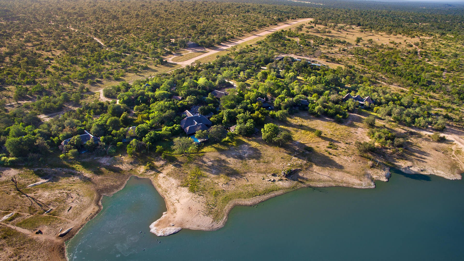 Aerial view of a lush green area with scattered buildings next to a body of water and surrounded by dry land and sparse vegetation. Paths and roads are visible, leading towards the horizon at Chitwa Chitwa a lodge perfect for a safari elopement