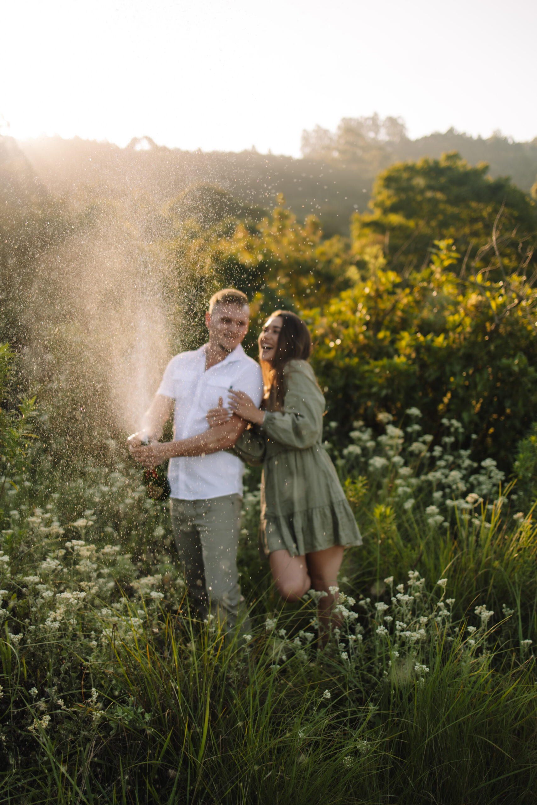 Engaged couple wearing cute engagements in a flower field popping champagne, perfect inspiration to help you on what to wear for your engagement photos