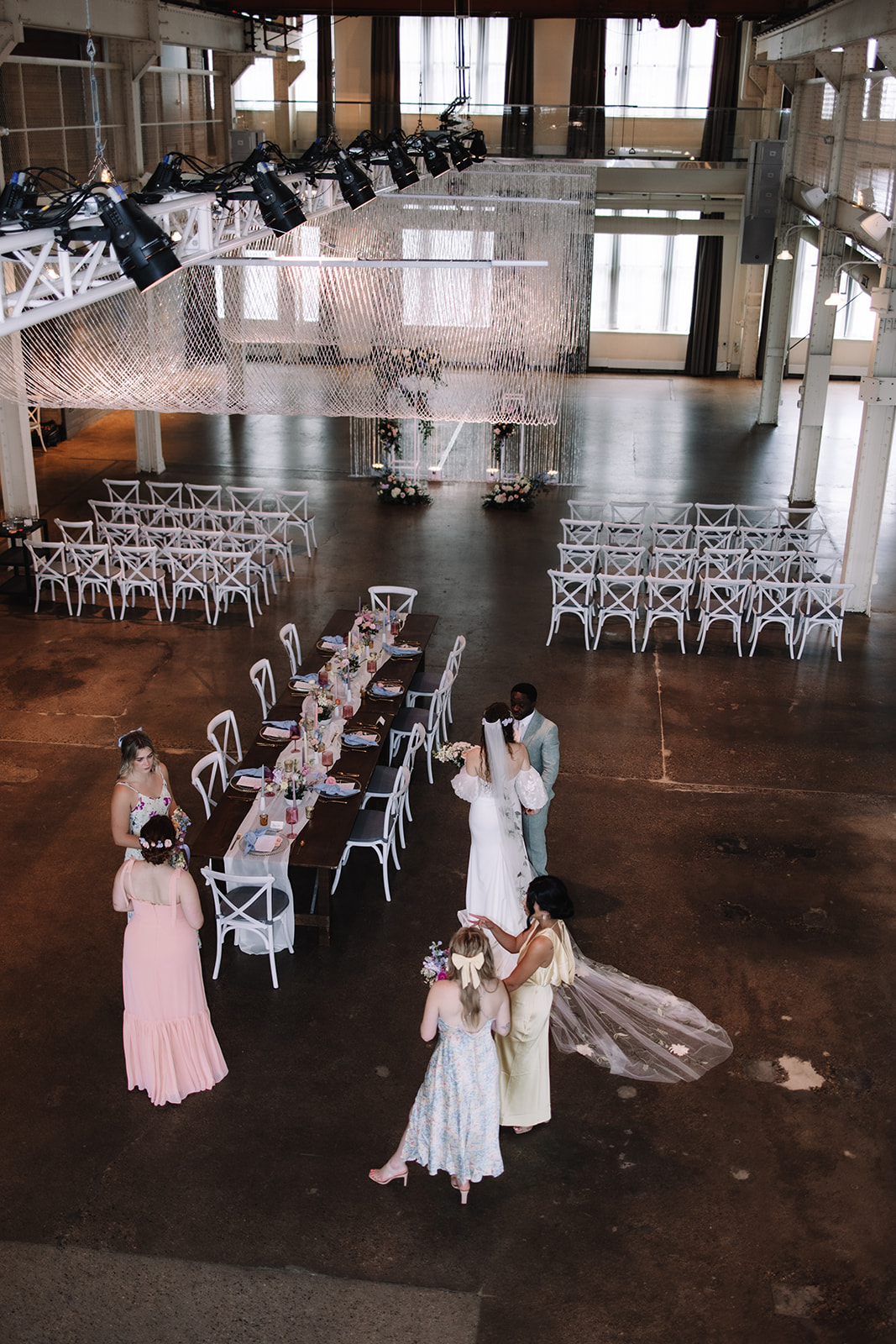 A wedding rehearsal in a spacious, industrial-style venue with large windows. Six people are gathered; the bride is dressed in white with a long veil, and the wedding party is wearing various pastel gowns and suits at the Machine Shop in Minneapolis