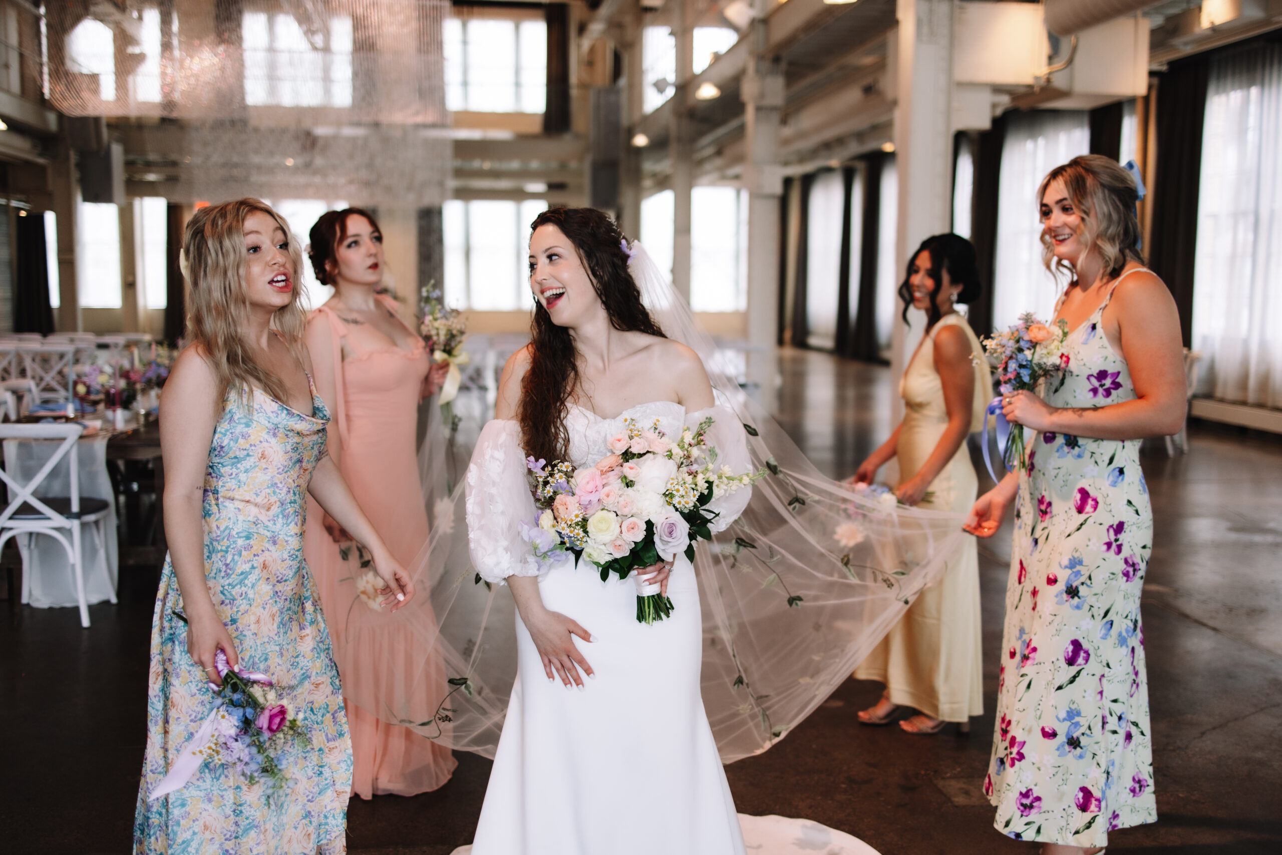 Bridesmaids dressed in mismatched, pastel, floral dresses, helping the bride fluff her veil while she laughs with them at the machine shop in Minneapolis