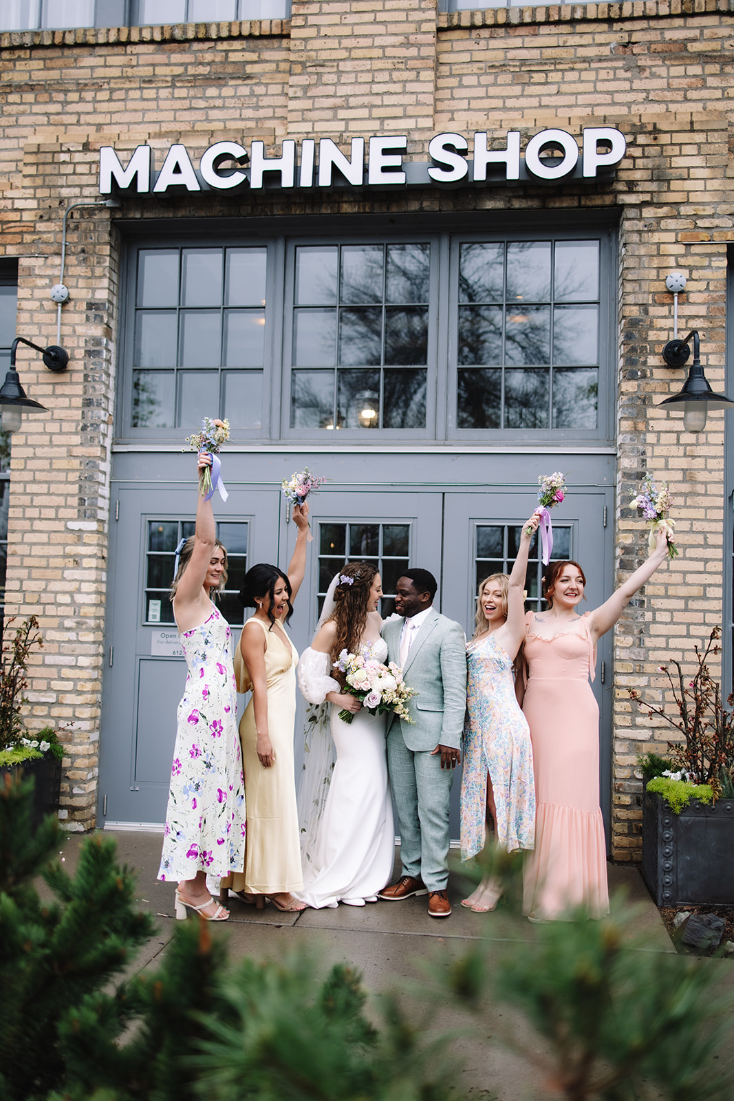 A wedding couple stands with their bridal party in front of a building labeled "Machine Shop." The group is dressed in formal attire, holding bouquets and smiling at each other.