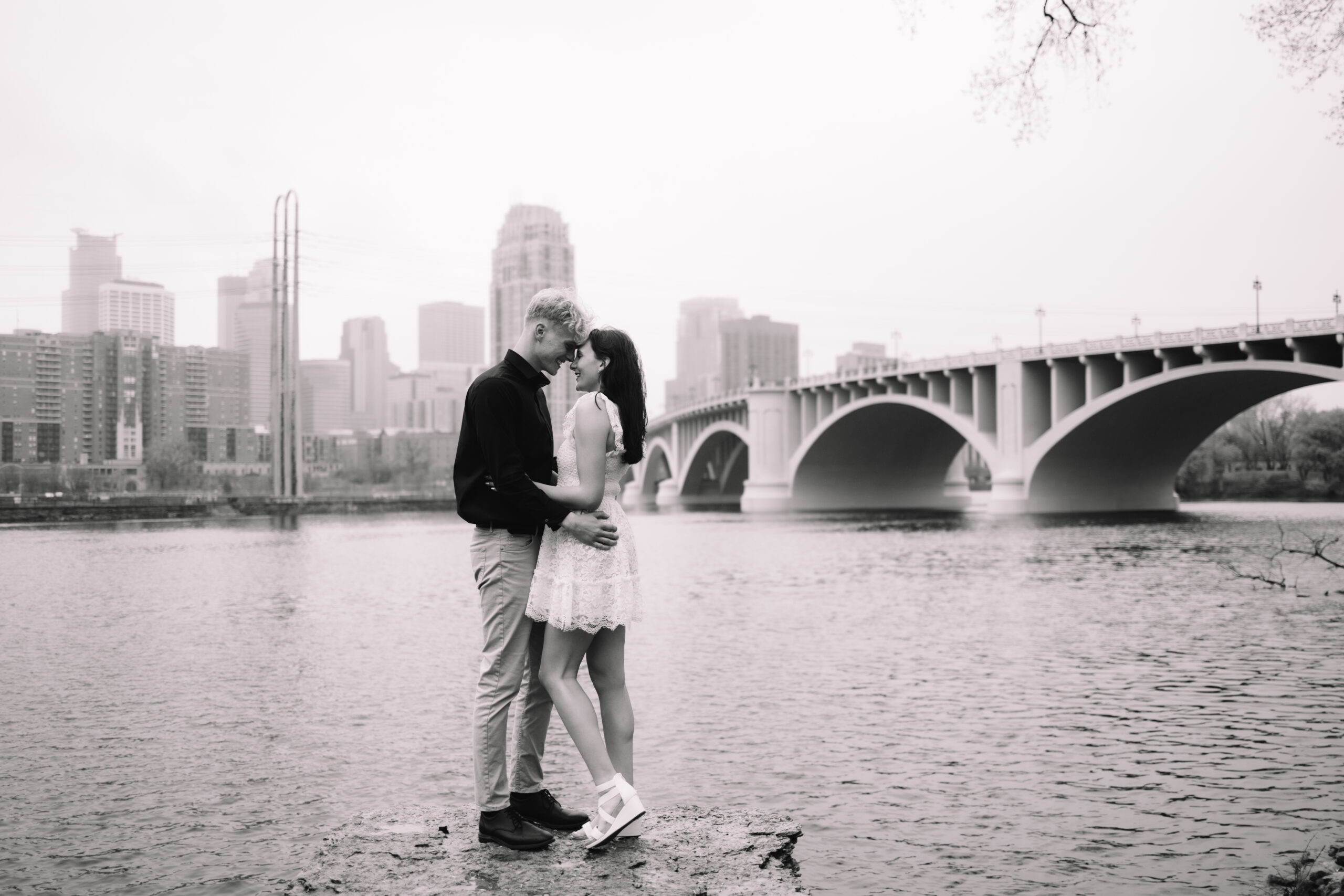 A couple stands on a rock by a river, embracing and kissing, with a city skyline and a bridge in the background.