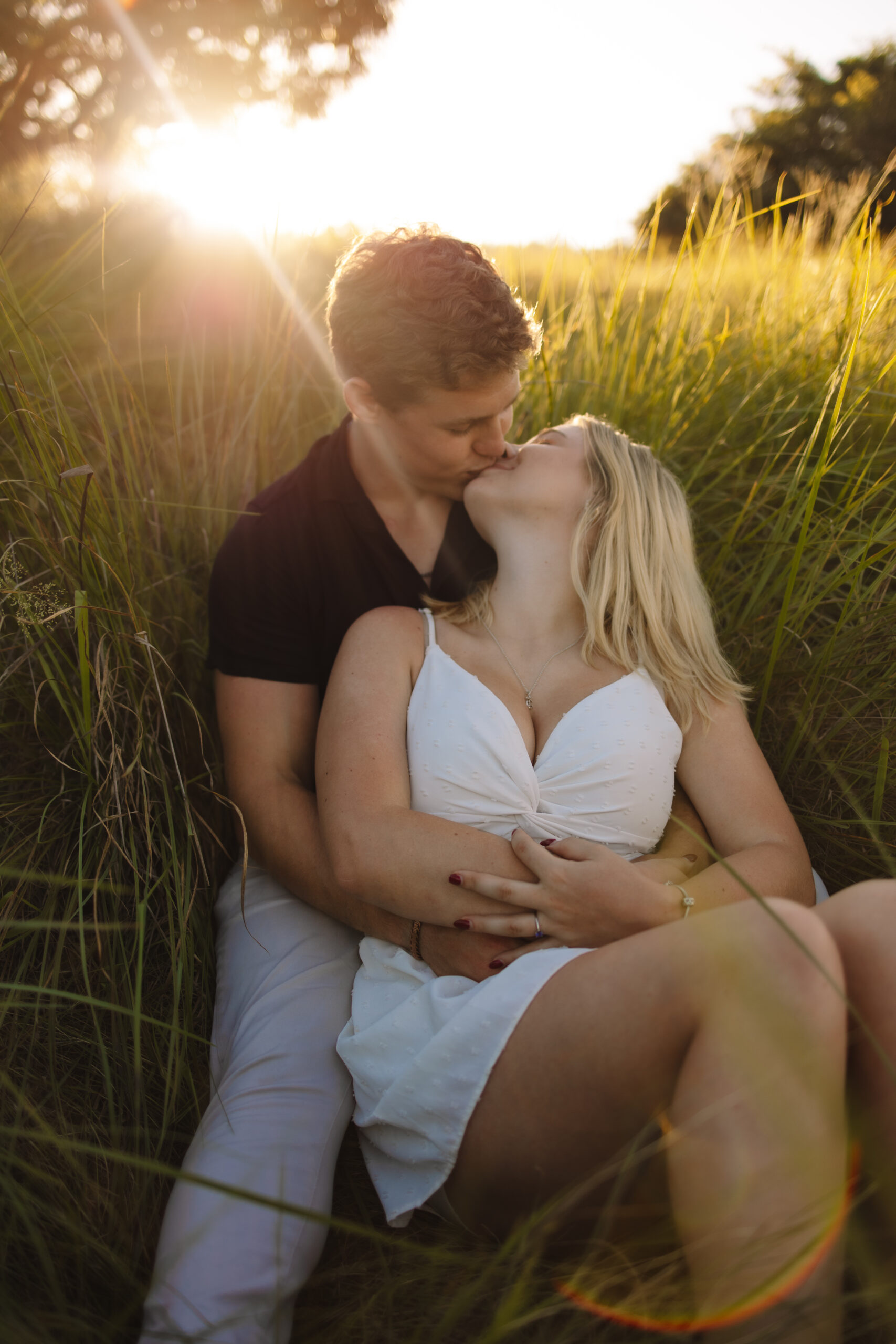 Newly engaged couple sitting in a field of grass kissing wearing casual and formal engagement outfits