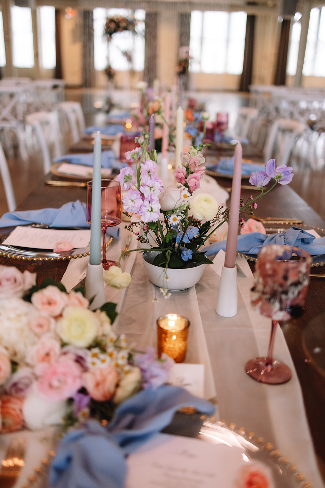 A long dining table is elegantly set for an event with white candles, pink and purple flowers, glassware, and place settings. The centerpiece includes a variety of flowers in a white bowl.