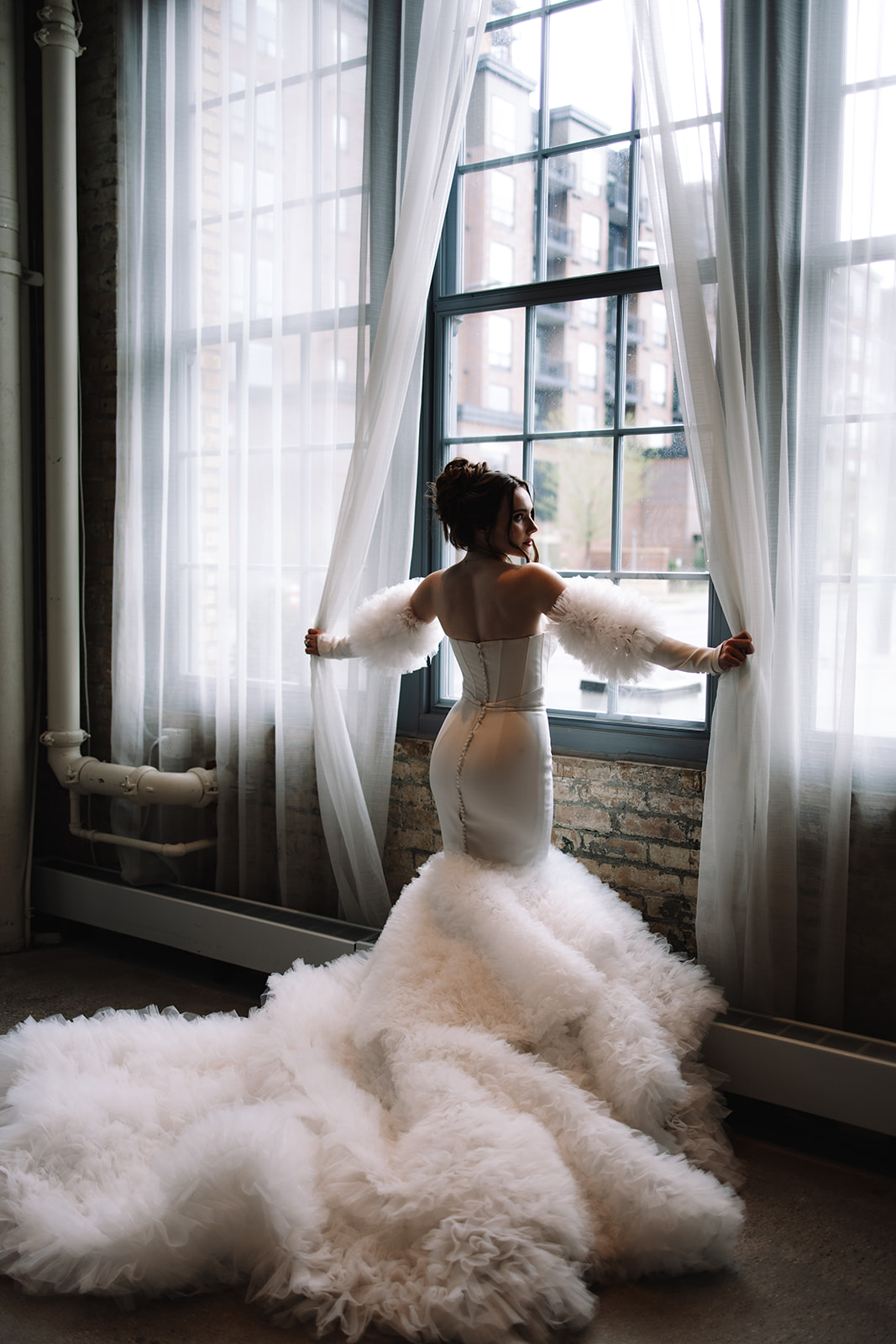 A woman in a strapless white dress with a mermaid silhouette and feathered sleeves stands by a large window, looking over her shoulder at the machine Shop in Minneapolis