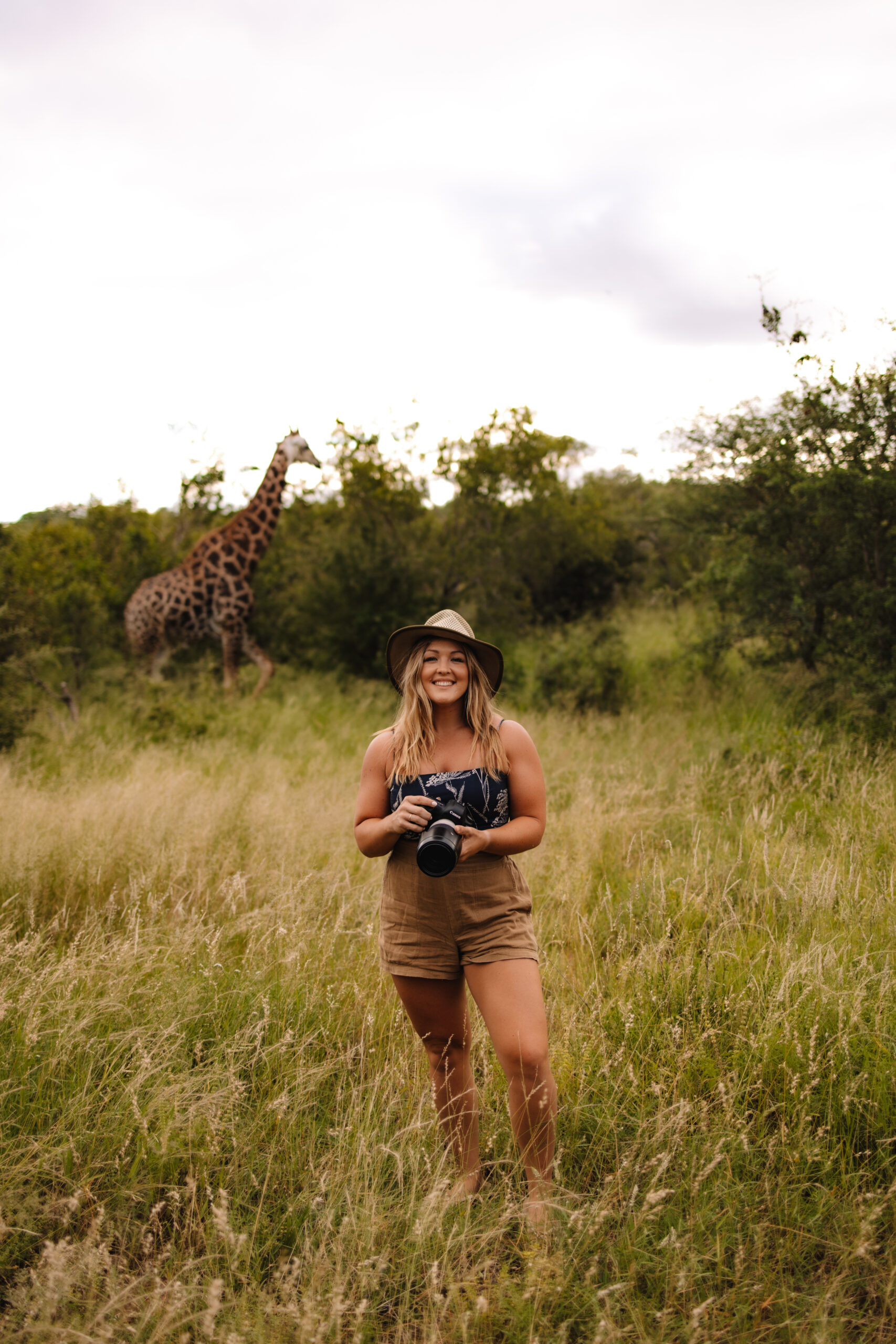 A wedding photographer with her camera standing in the long green grass with a giraffe walking behind her